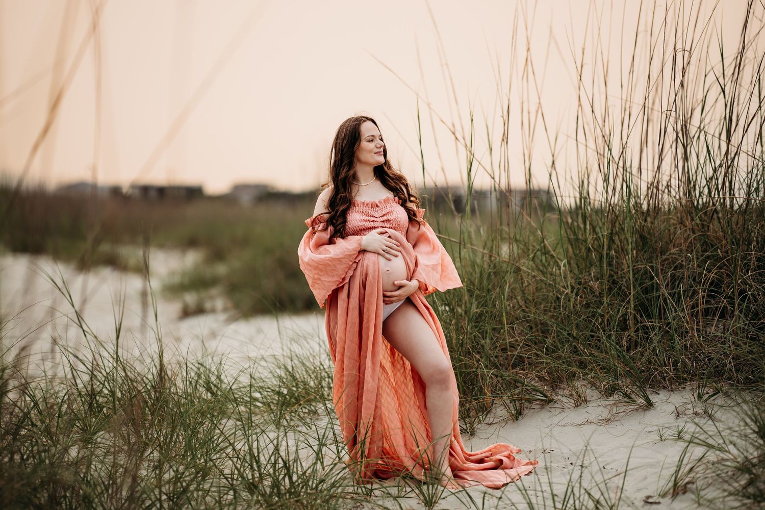 Pregnant woman in flowing coral maternity dress posing among sea oats on sandy beach at sunset.