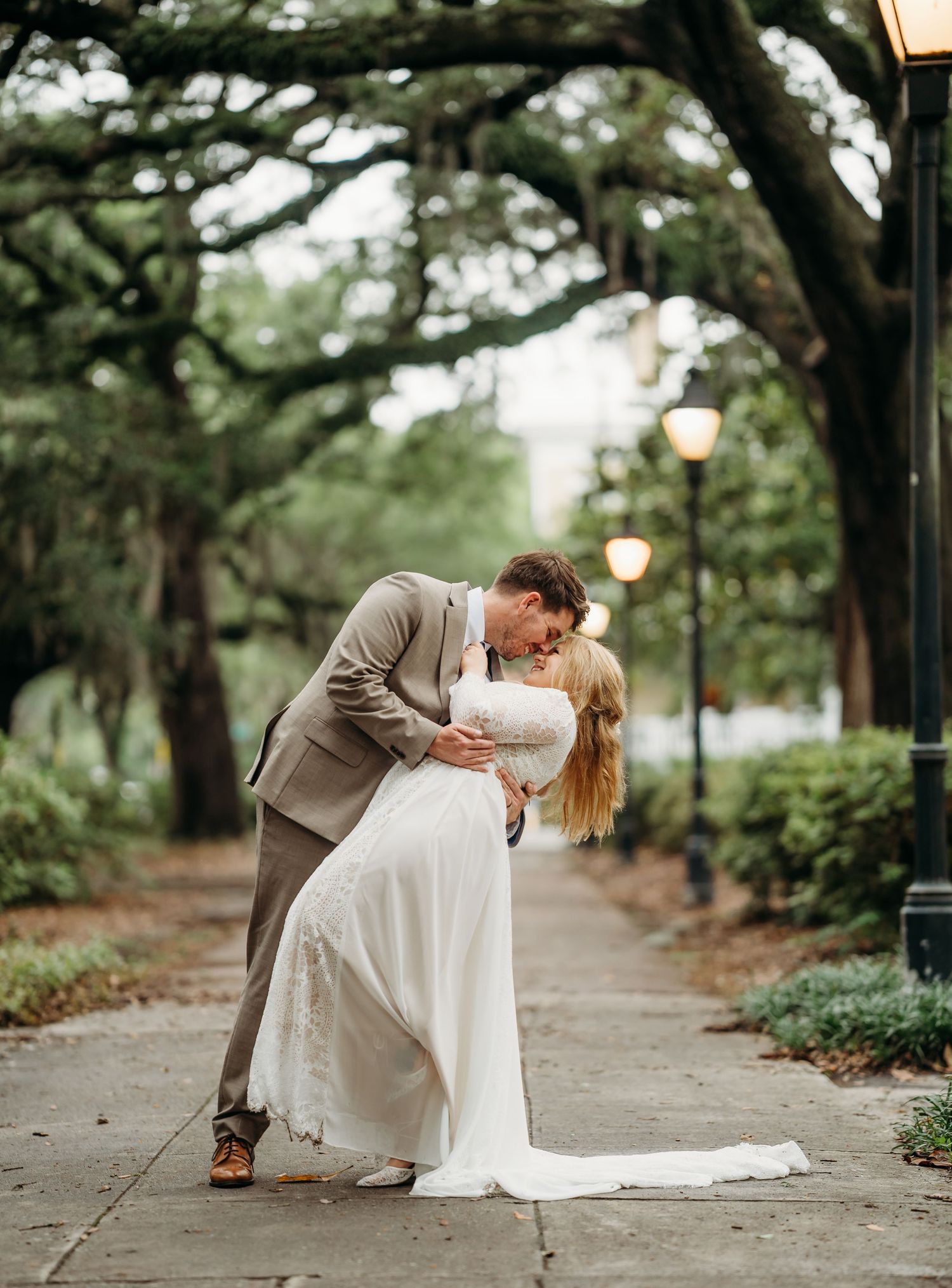 Groom dips bride in a romantic kiss on a scenic tree-lined pathway glowing with vintage lanterns at twilight.