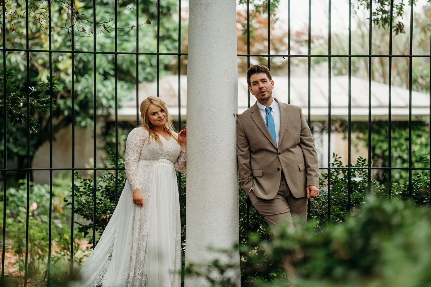 Bride and groom posing on opposite sides of a white column surrounded by lush greenery at an outdoor wedding.