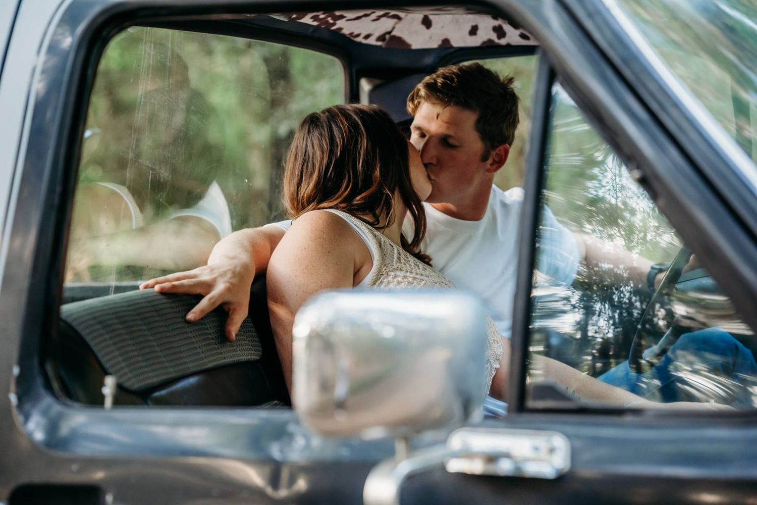 Romantic couple embracing inside vintage truck viewed through window.