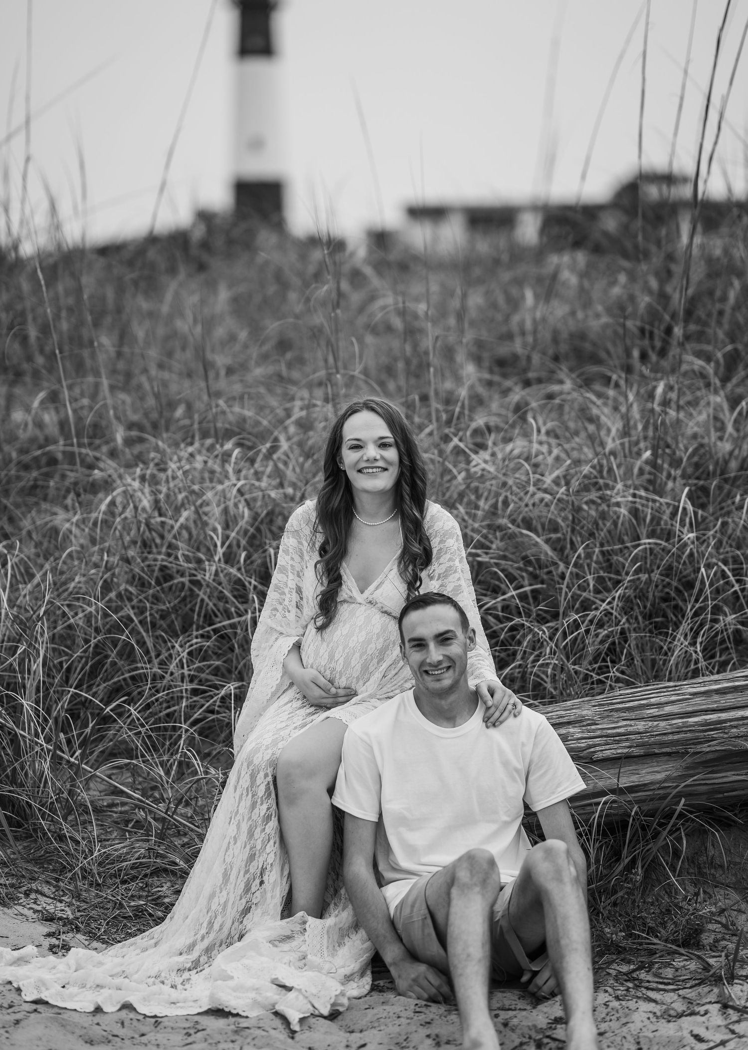 Black and white maternity photo of a couple sitting on driftwood at beach with lighthouse backdrop.