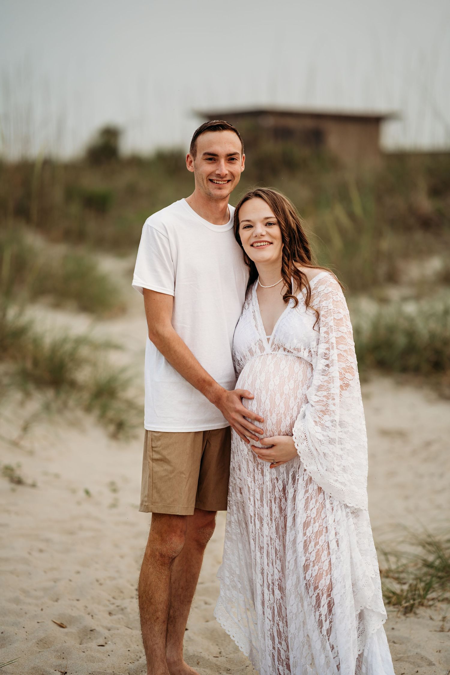 Smiling couple posing together on beach, woman in white lace maternity dress holding baby bump.