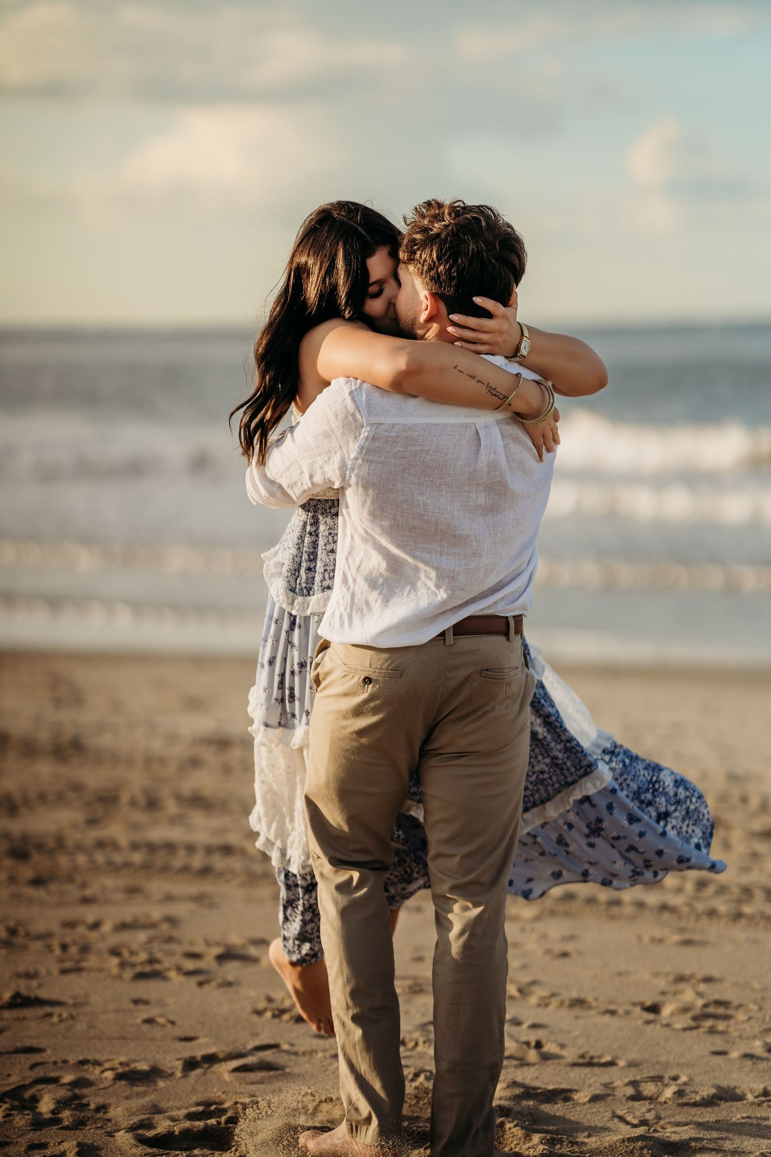 Romantic couple embracing and kissing on a sandy beach with ocean waves in the background.