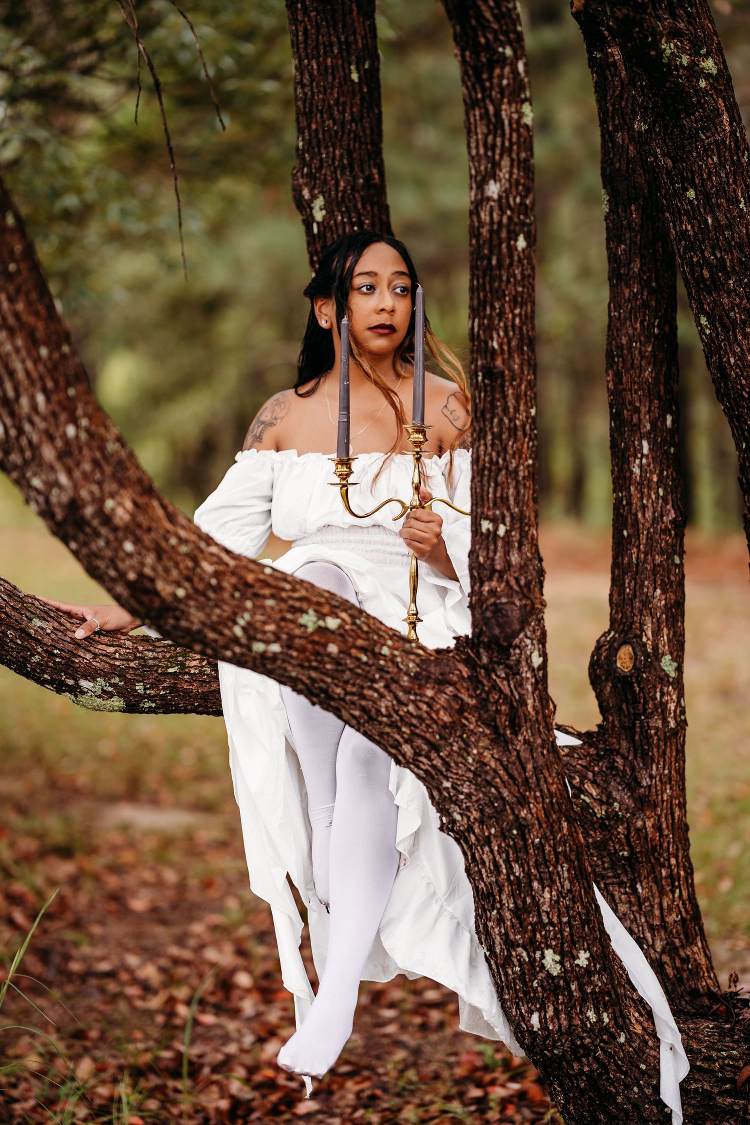 Woman in white off-shoulder dress leaning against tree trunk in forest.