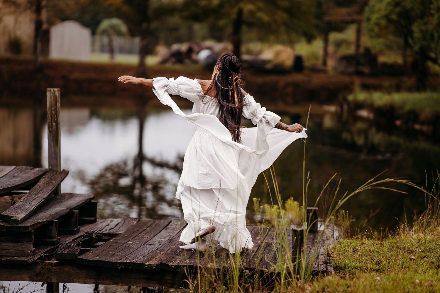 Woman in white dress twirls dramatically on rustic wooden dock beside tranquil water.