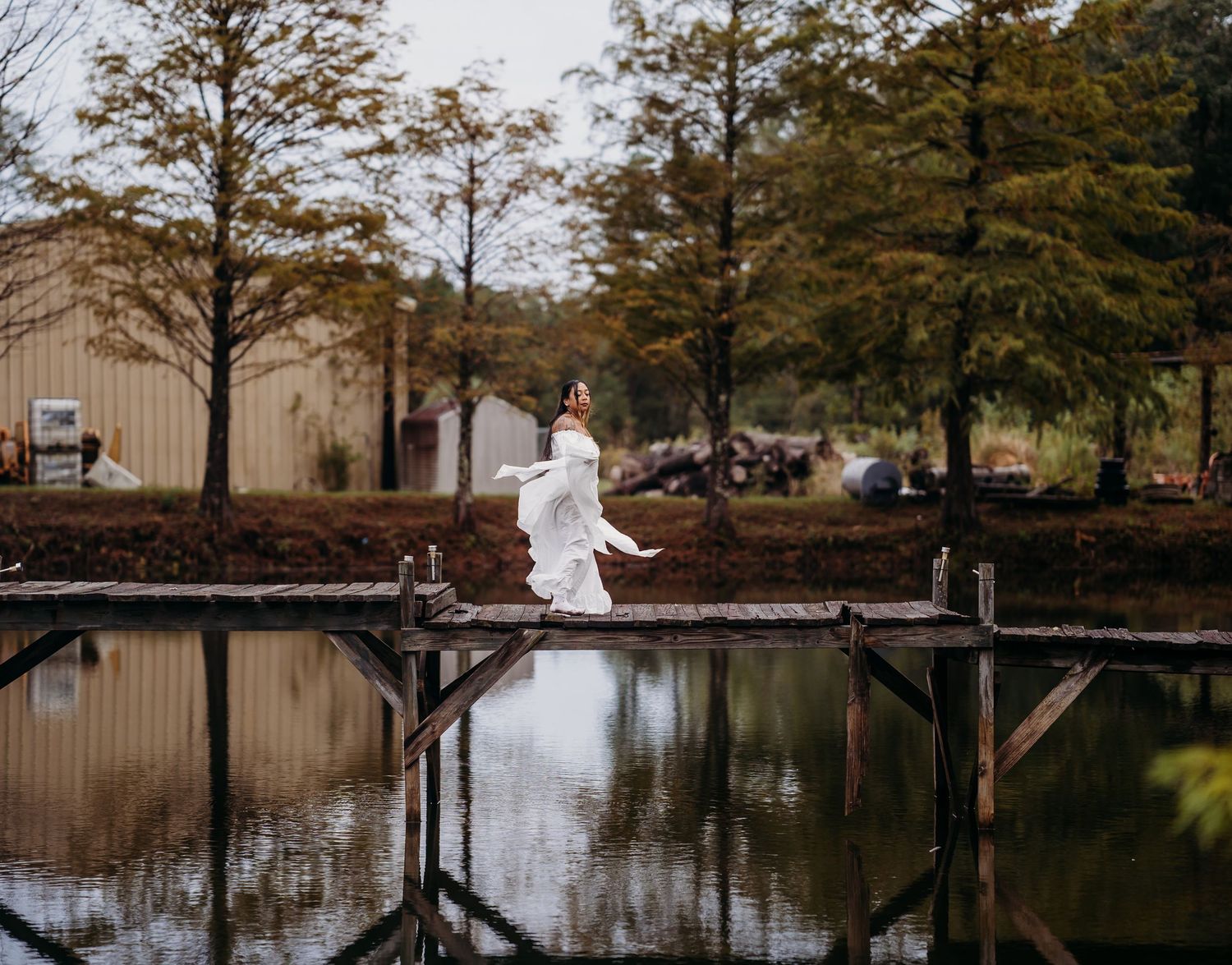 Bride in white wedding dress walking gracefully on wooden dock with pond reflection and fall foliage backdrop.