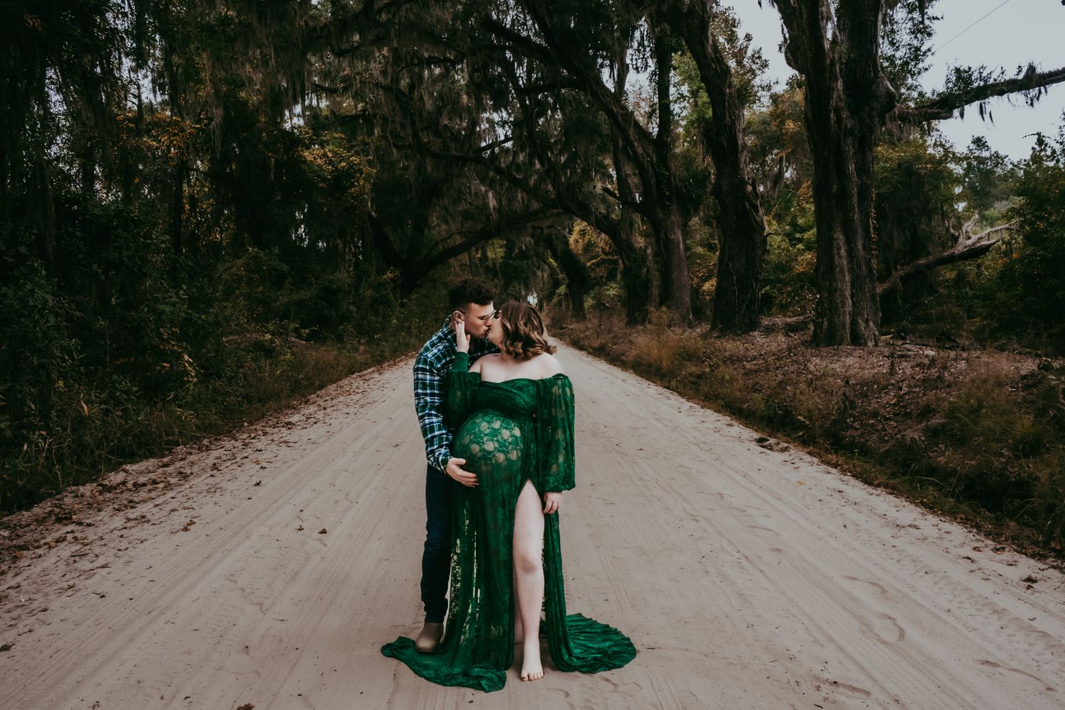Couple embracing on a sandy road with woman in elegant green maternity gown under oak trees.