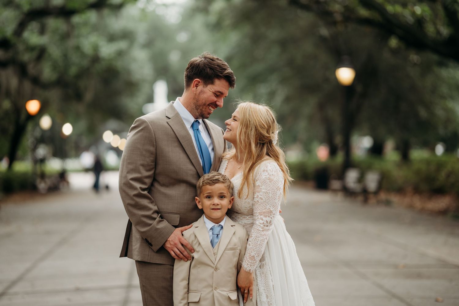 Bride and groom share tender moment with young boy between them on tree-lined Forsyth Park path.