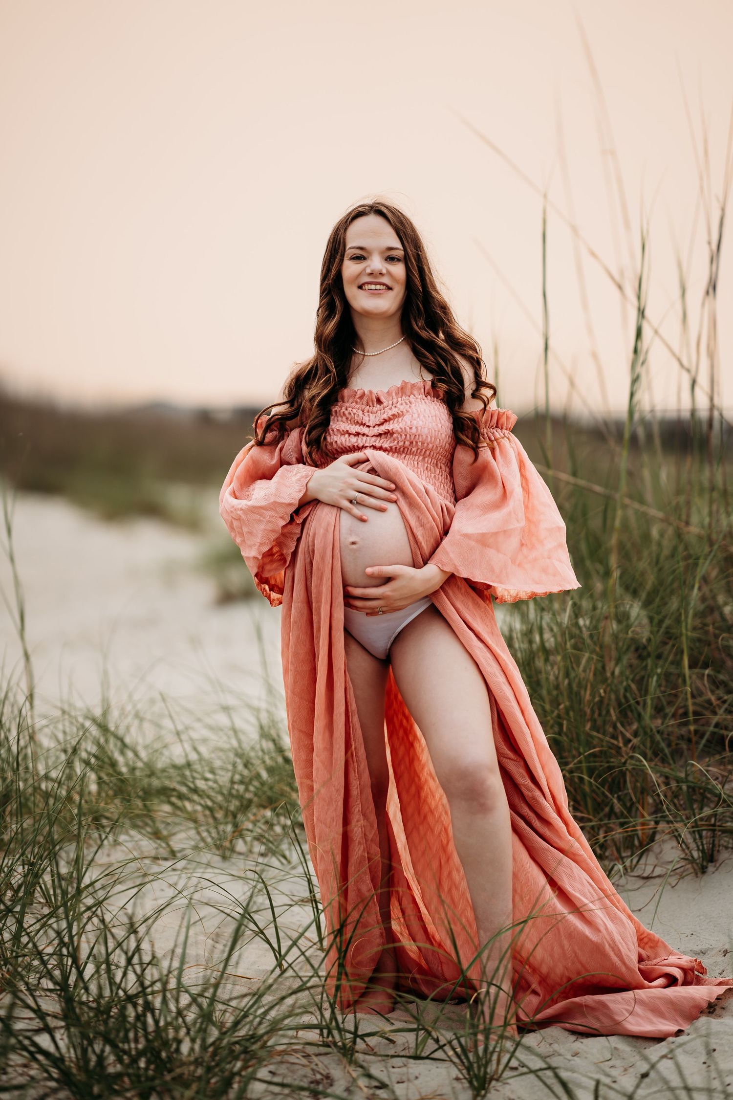 Radiant pregnant woman in coral maternity gown standing on beach dunes surrounded by sea grass.