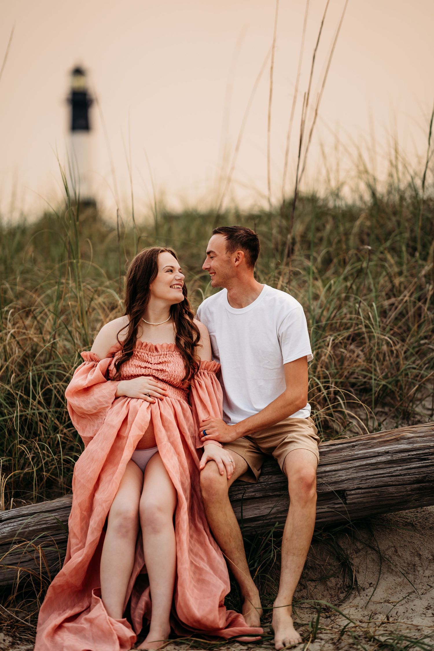 Expecting couple sitting on driftwood smiling at each other near lighthouse during beach session.