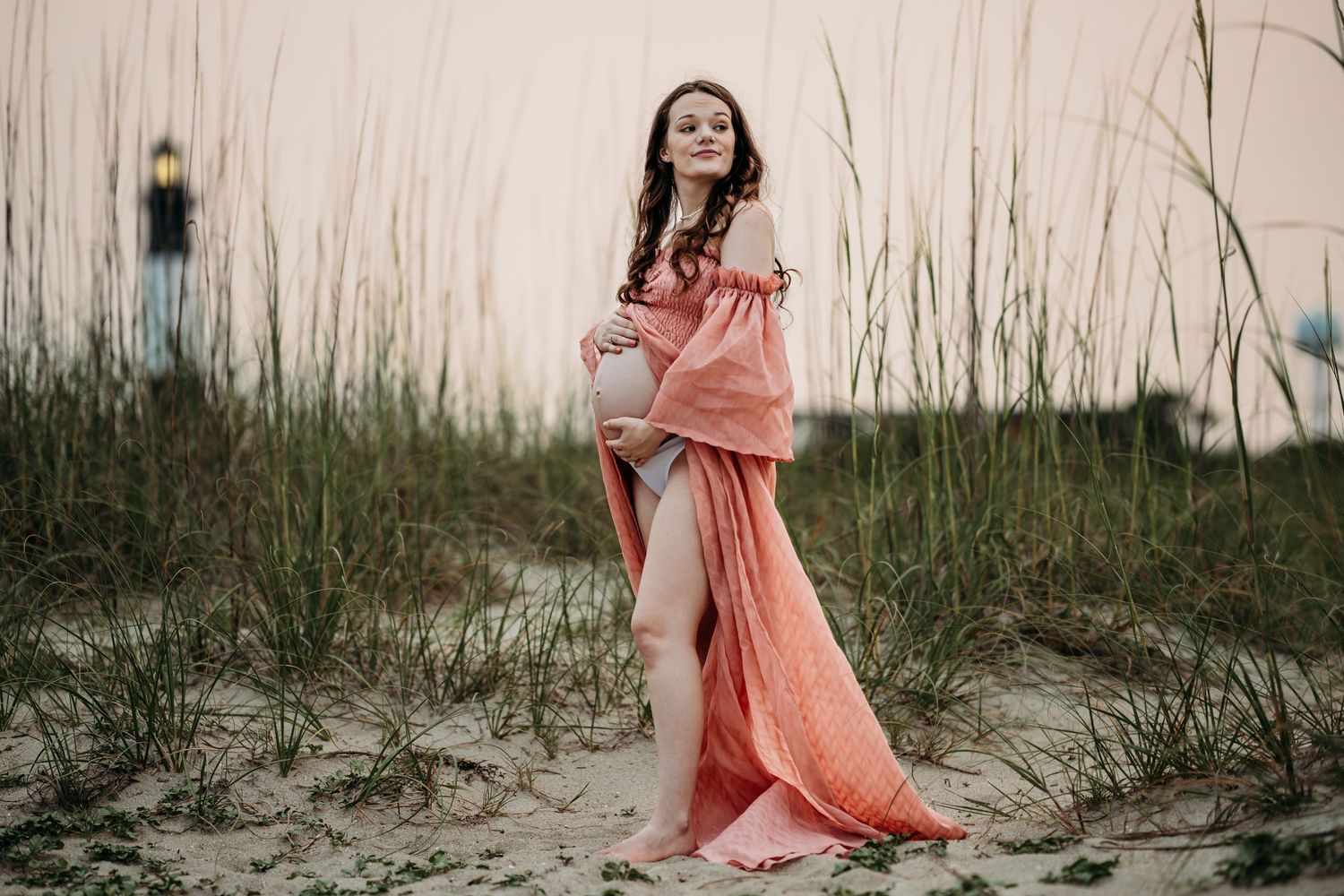 Glowing pregnant woman in flowing coral off-shoulder dress poses in sandy beach dunes at sunset.