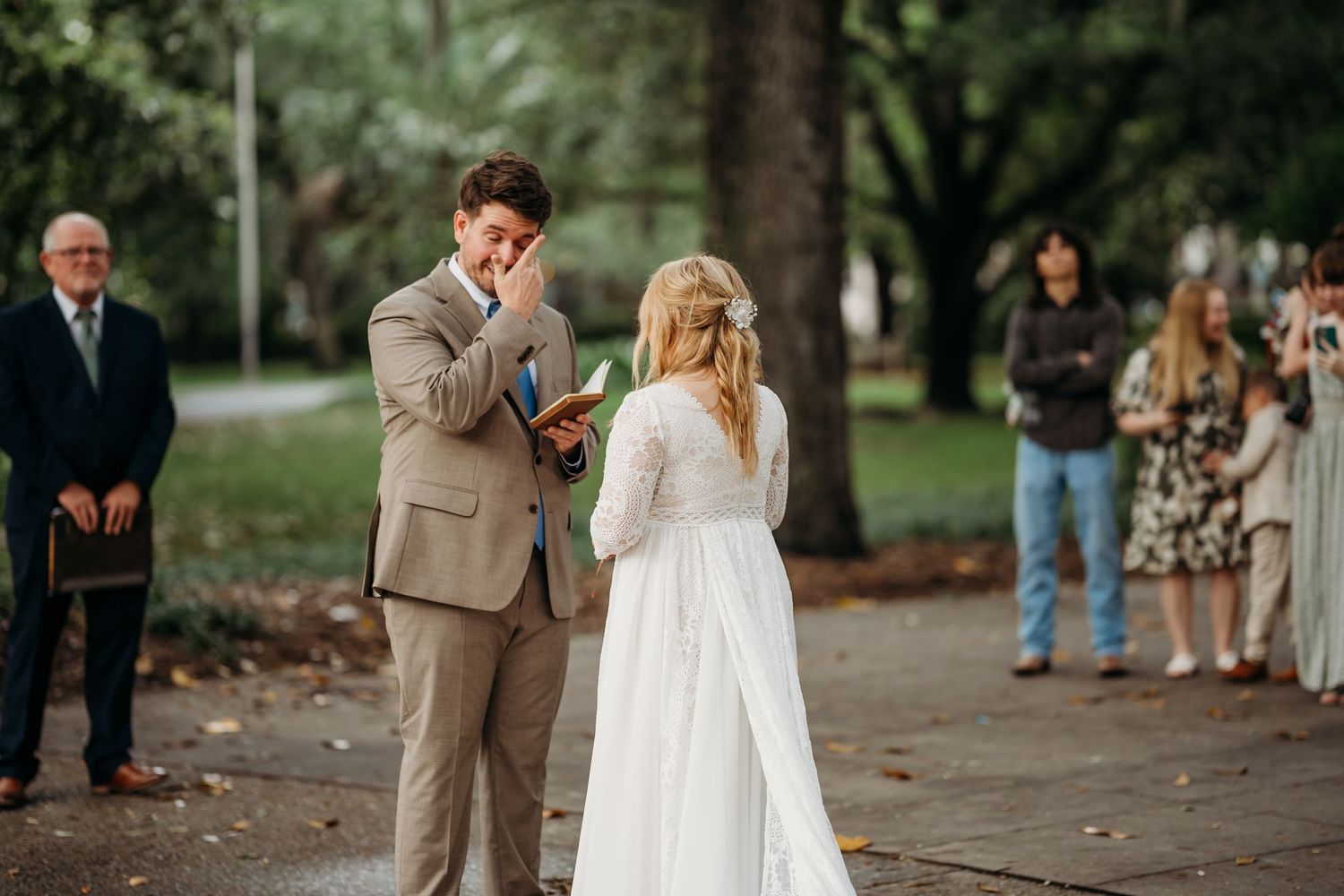 Emotional groom wipes tears while reading vows to bride during outdoor park wedding ceremony.