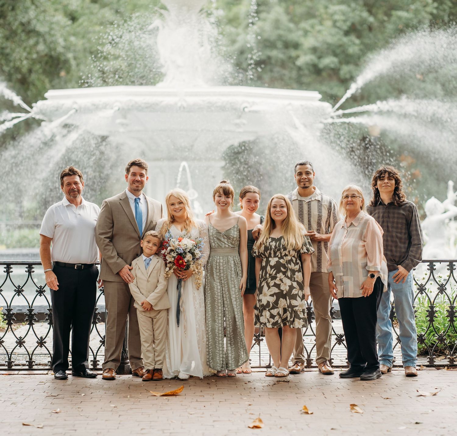 Wedding party group photo with bride, groom, and family posing before a large white fountain.