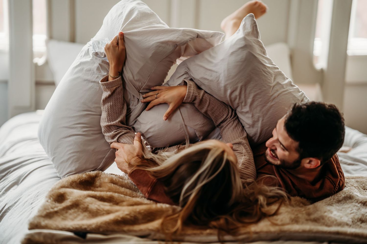 Playful couple having a pillow fight while laughing together on a cozy bed.