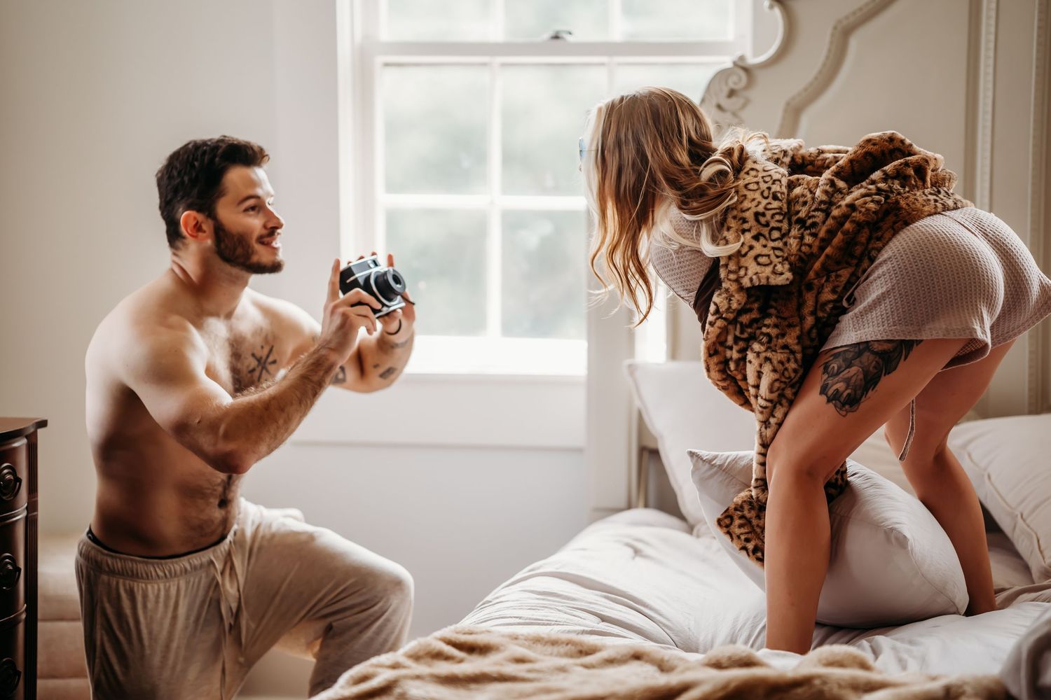 Shirtless tattooed man photographing woman climbing onto white ornate bed.