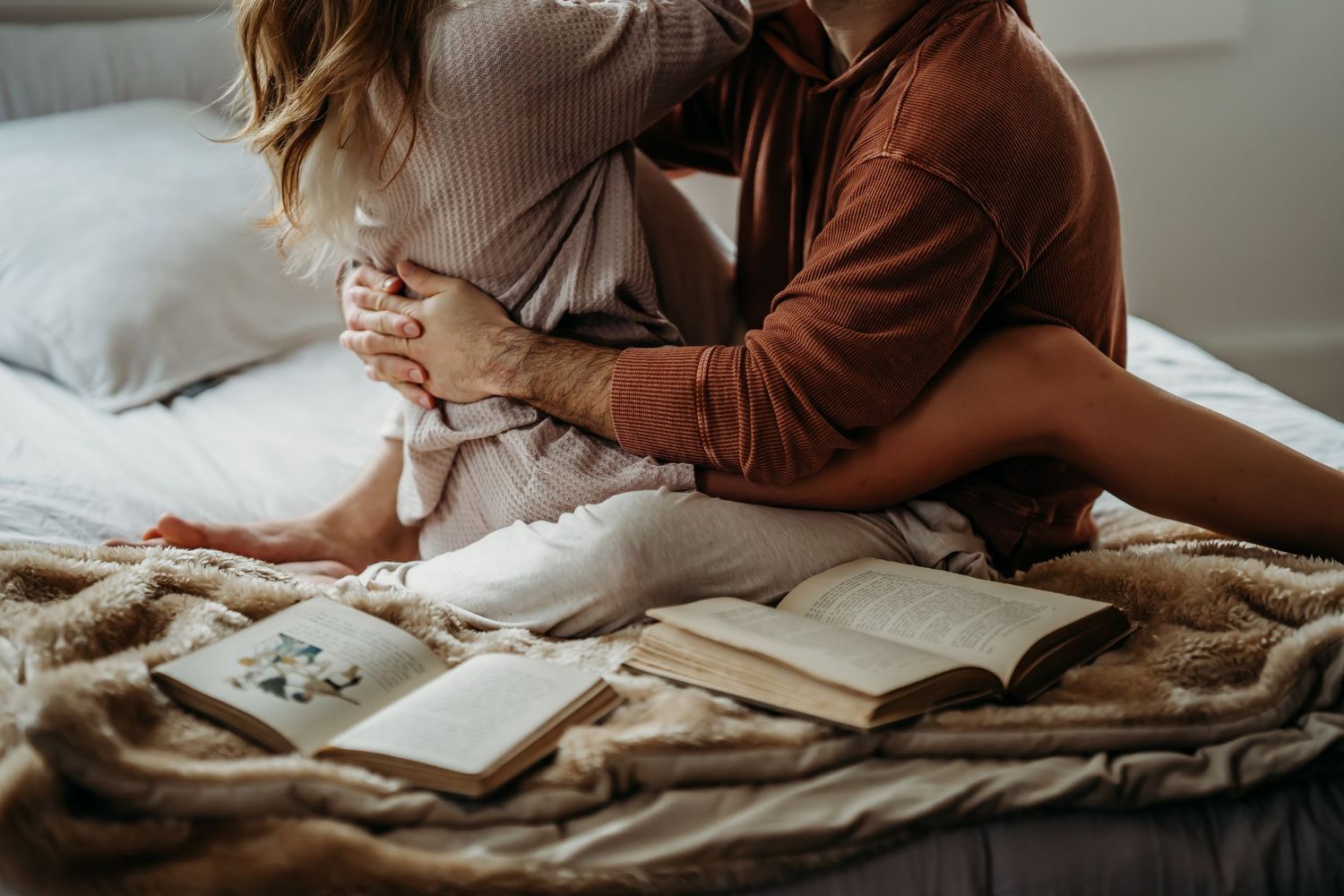 A couple sits intimately on a bed surrounded by open books on a soft beige blanket.