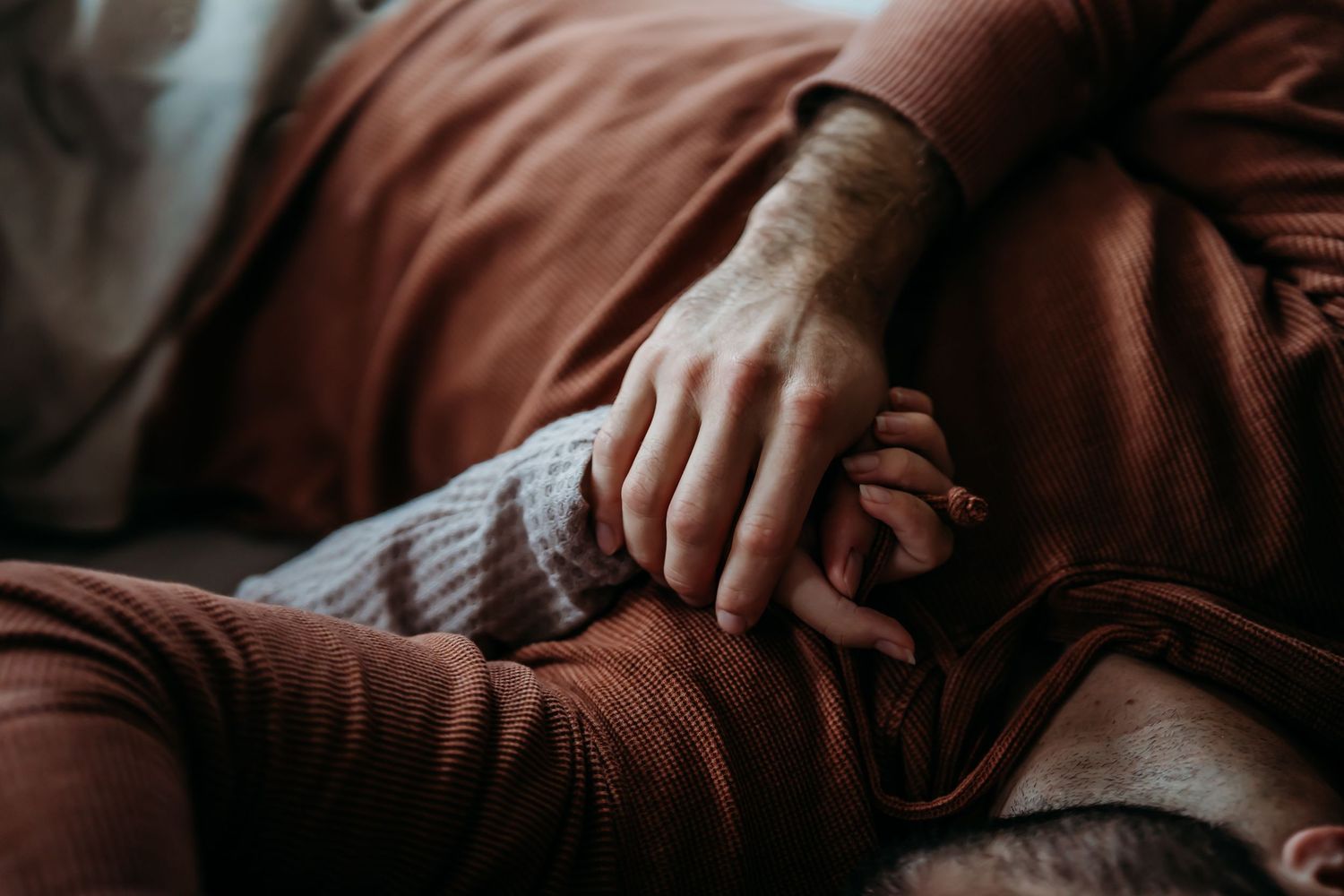 Close-up of couple holding hands tenderly on bed with warm brown bedding.