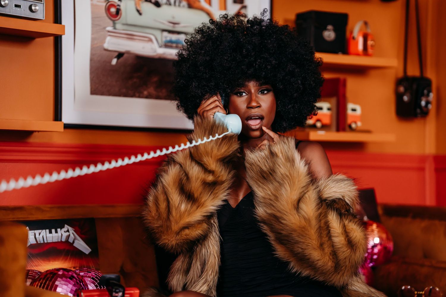 Stylish woman with afro hairstyle in fur coat talks on retro phone in vibrant orange room.