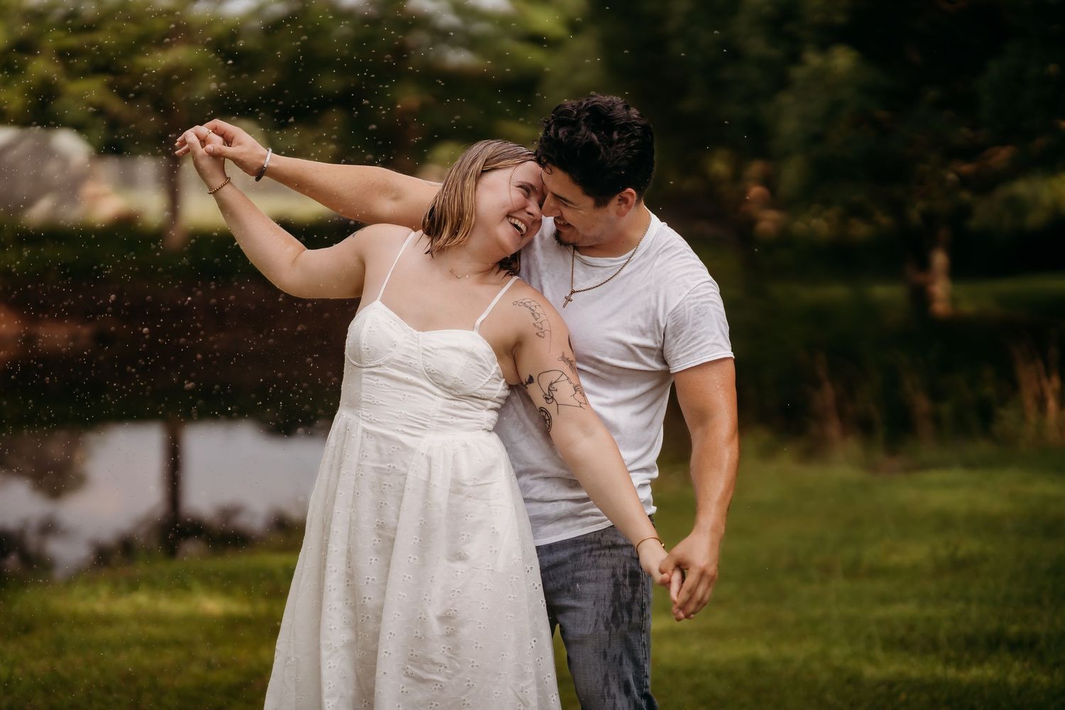 Couple dancing outdoors in the rain, woman in white dress and man in white t-shirt sharing a romantic moment in a green park.