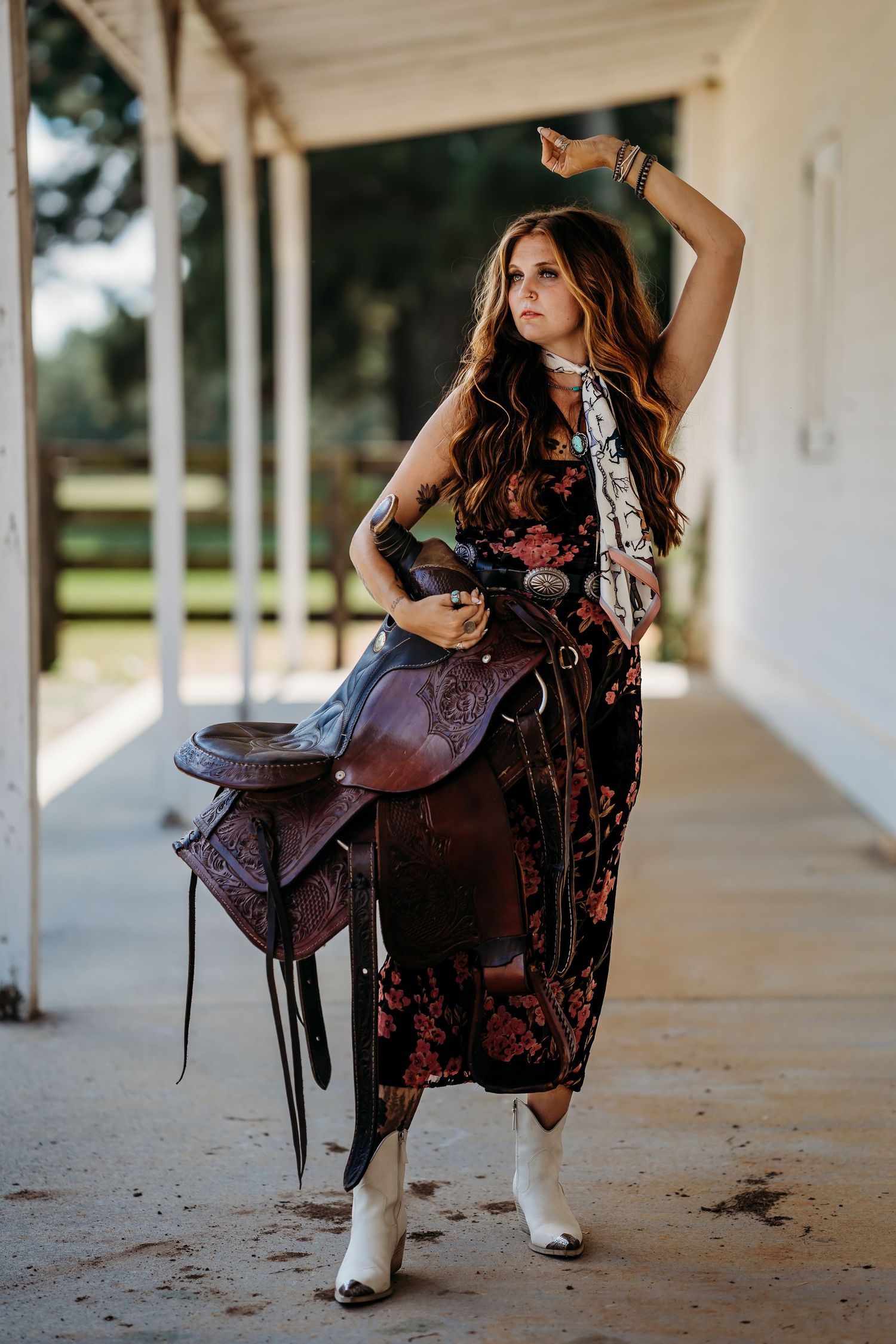 Woman in floral dress and white cowboy boots holding a leather saddle in a rustic barn setting.