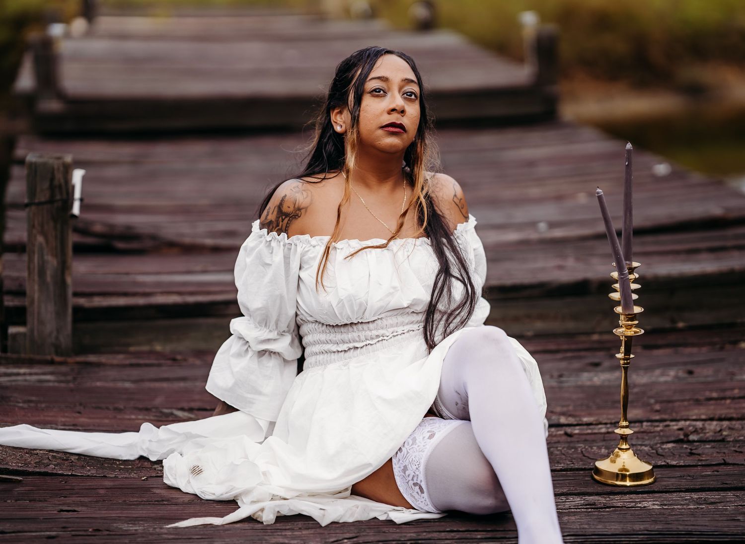 Woman in white off-shoulder dress and white thigh-high stockings sitting on wooden dock with gold candlestick.