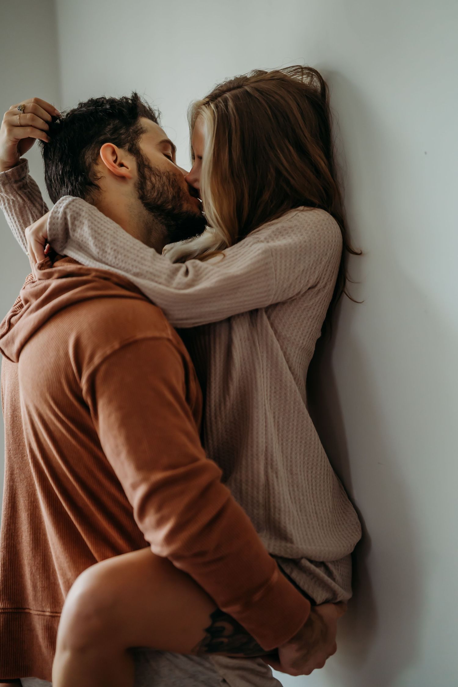 A couple sharing a passionate kiss against a white wall, the man in an orange hoodie lifting the woman in a beige sweater.