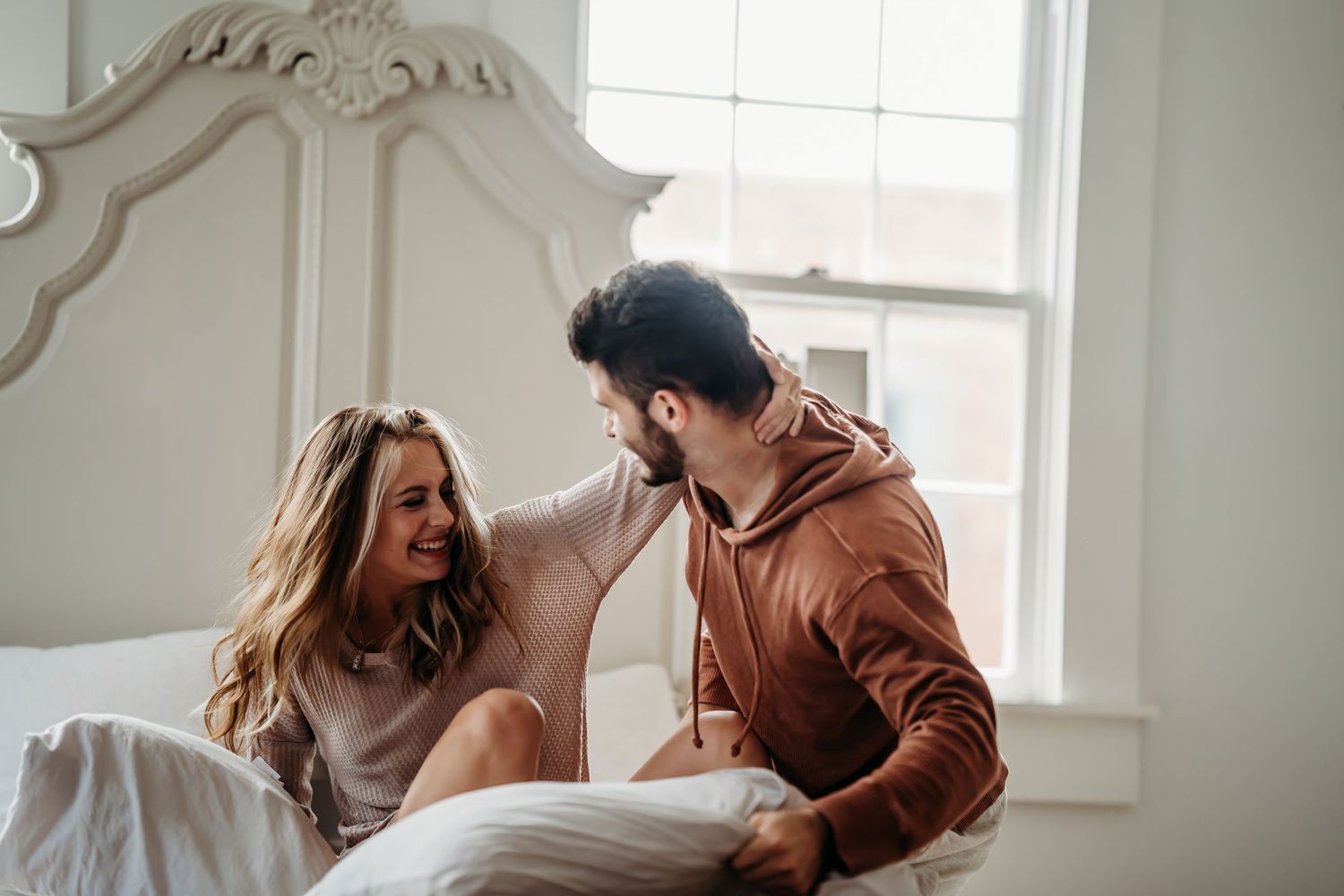 A happy couple sitting on a white bed, smiling and laughing together in a bright, cozy bedroom.
