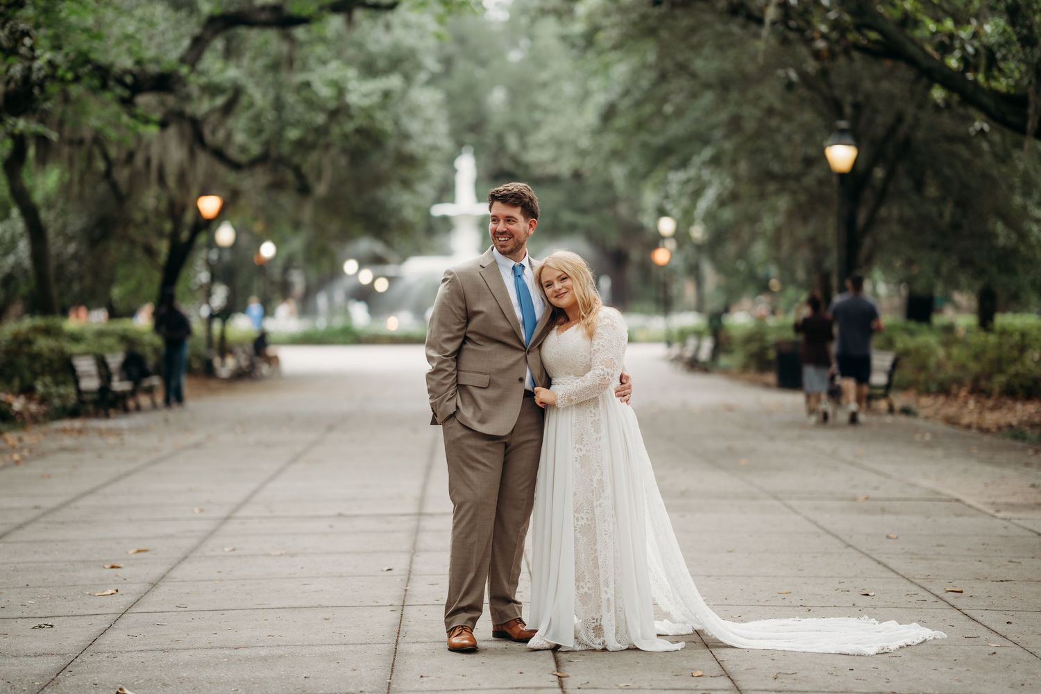 Bride and groom pose on a tree-lined walkway with a fountain backdrop in Forsyth Park, Savannah.