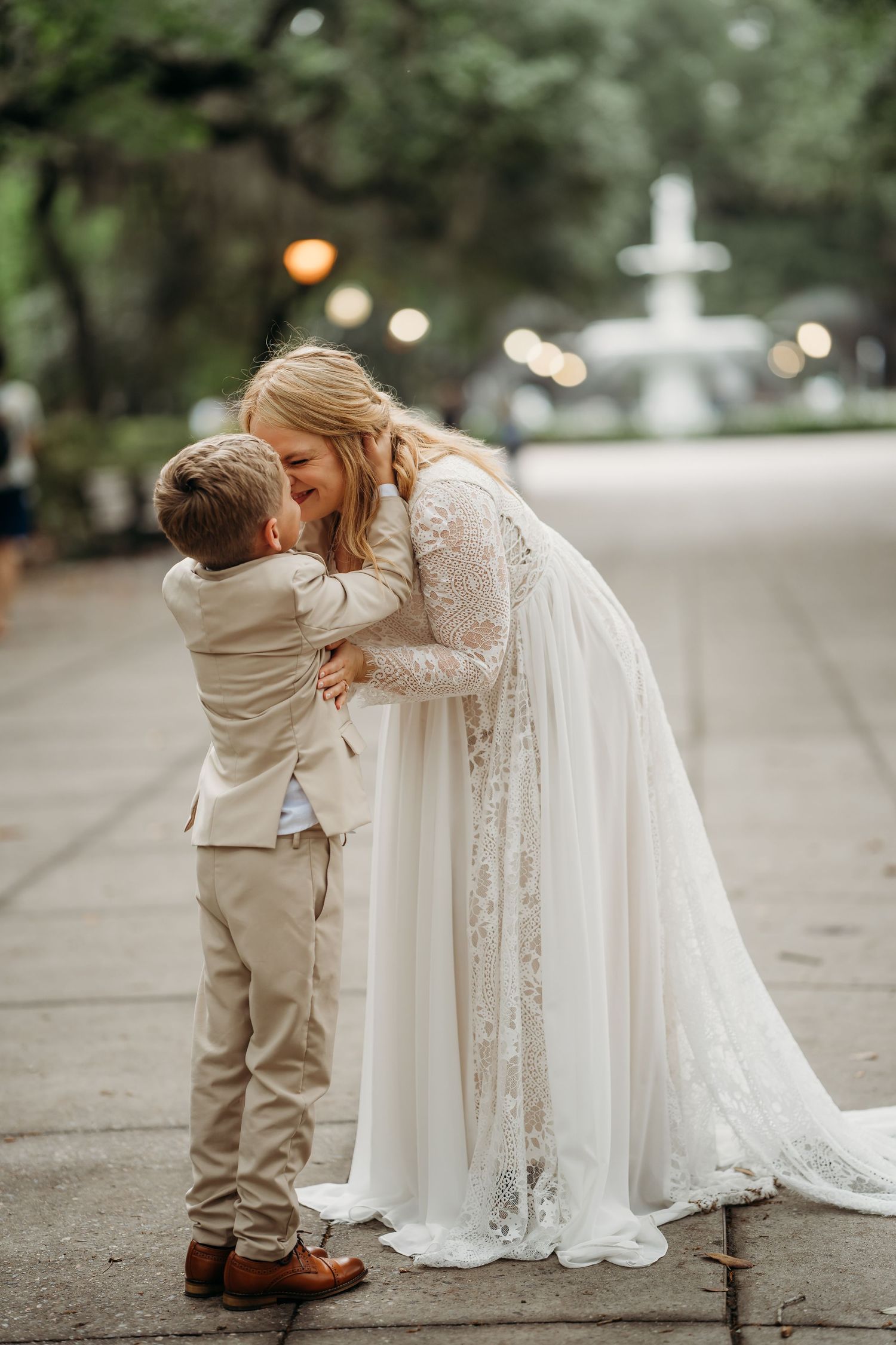 Bride in lace gown shares tender embrace with young boy in tan suit at park.