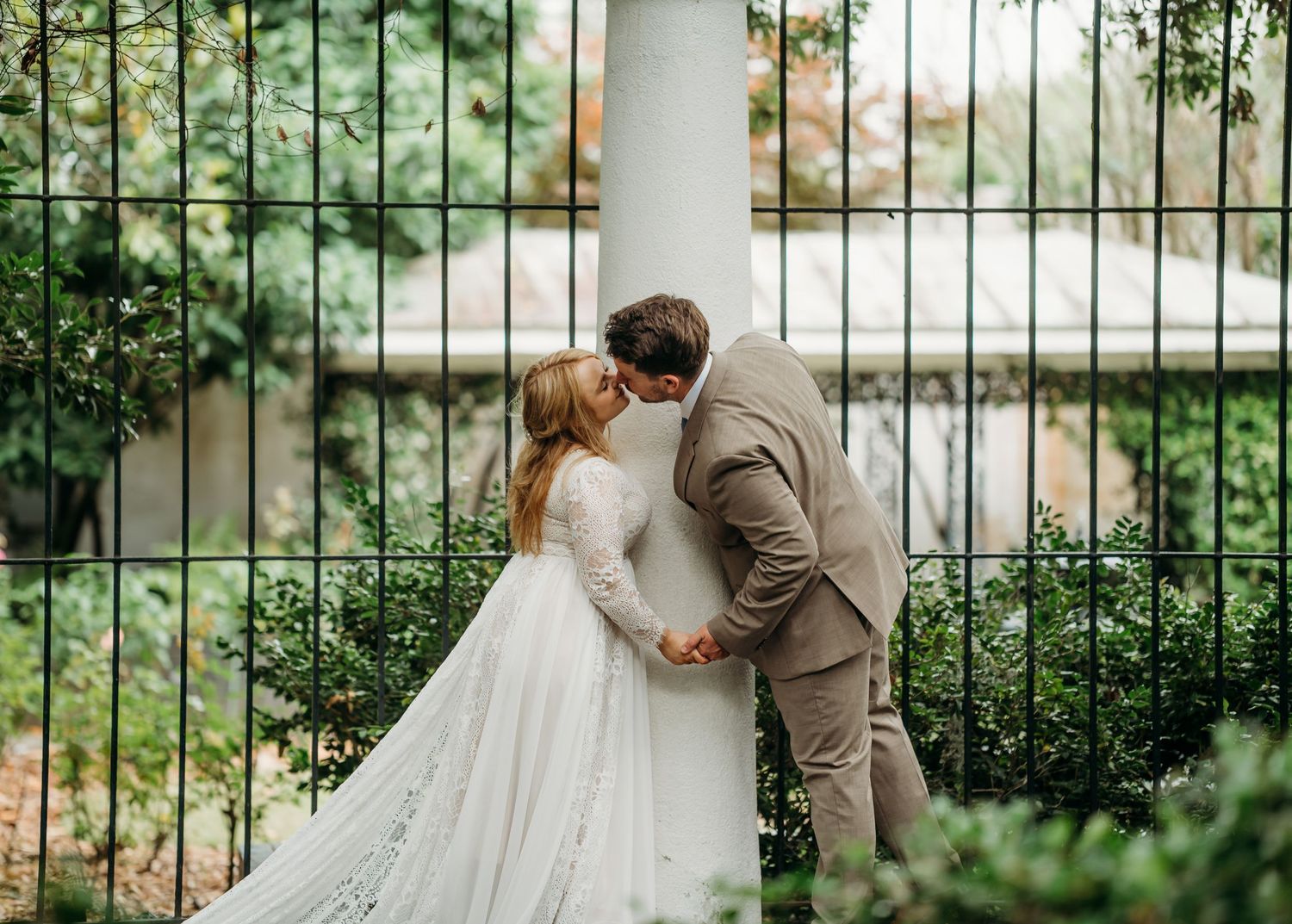 Bride and groom sharing a romantic kiss behind a white column with a lush green garden fence background.