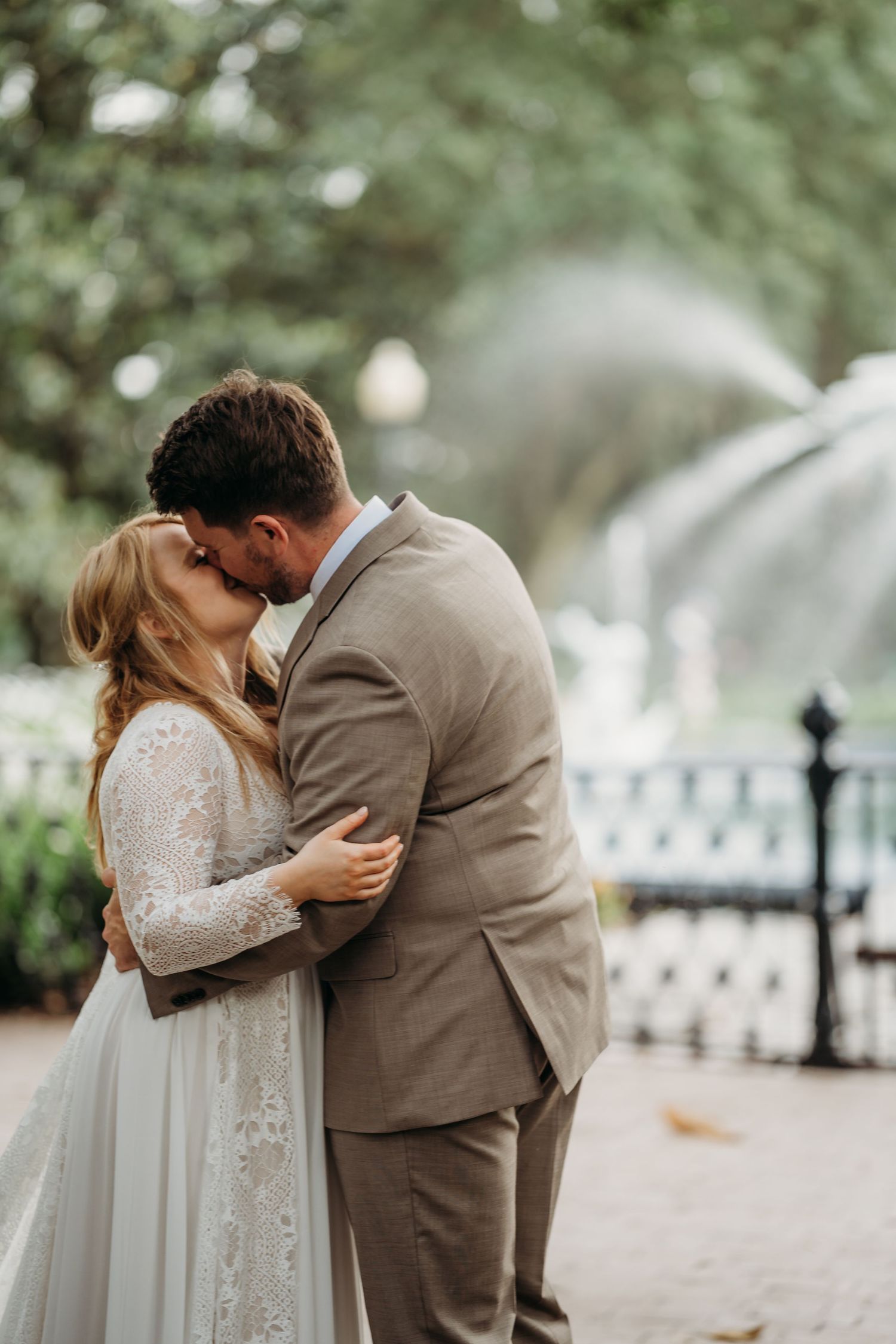 Newlyweds kiss tenderly in front of a softly blurred fountain backdrop on their wedding day.