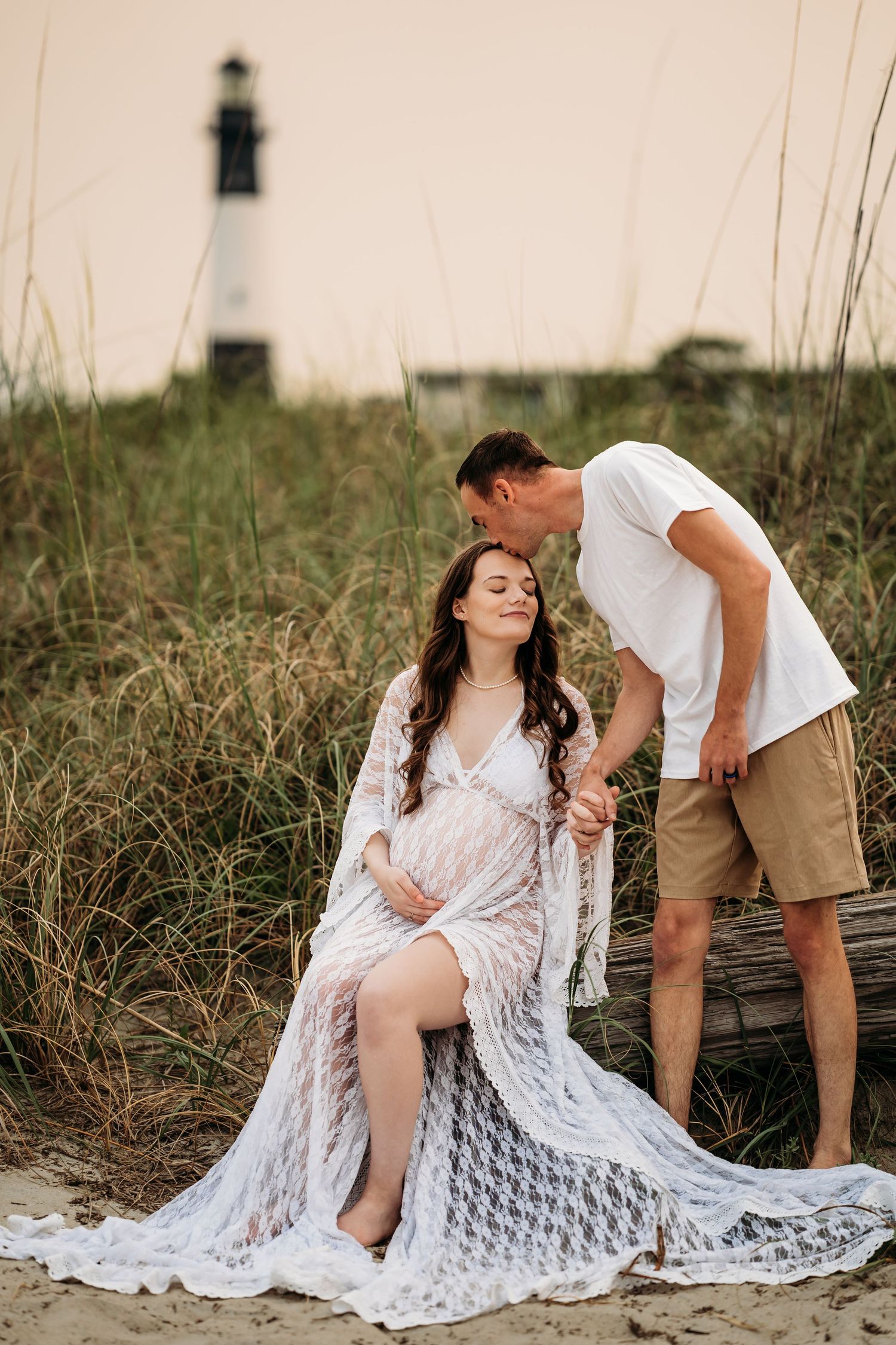 Couple posing near lighthouse at sunset, man kissing pregnant woman in white lace gown.