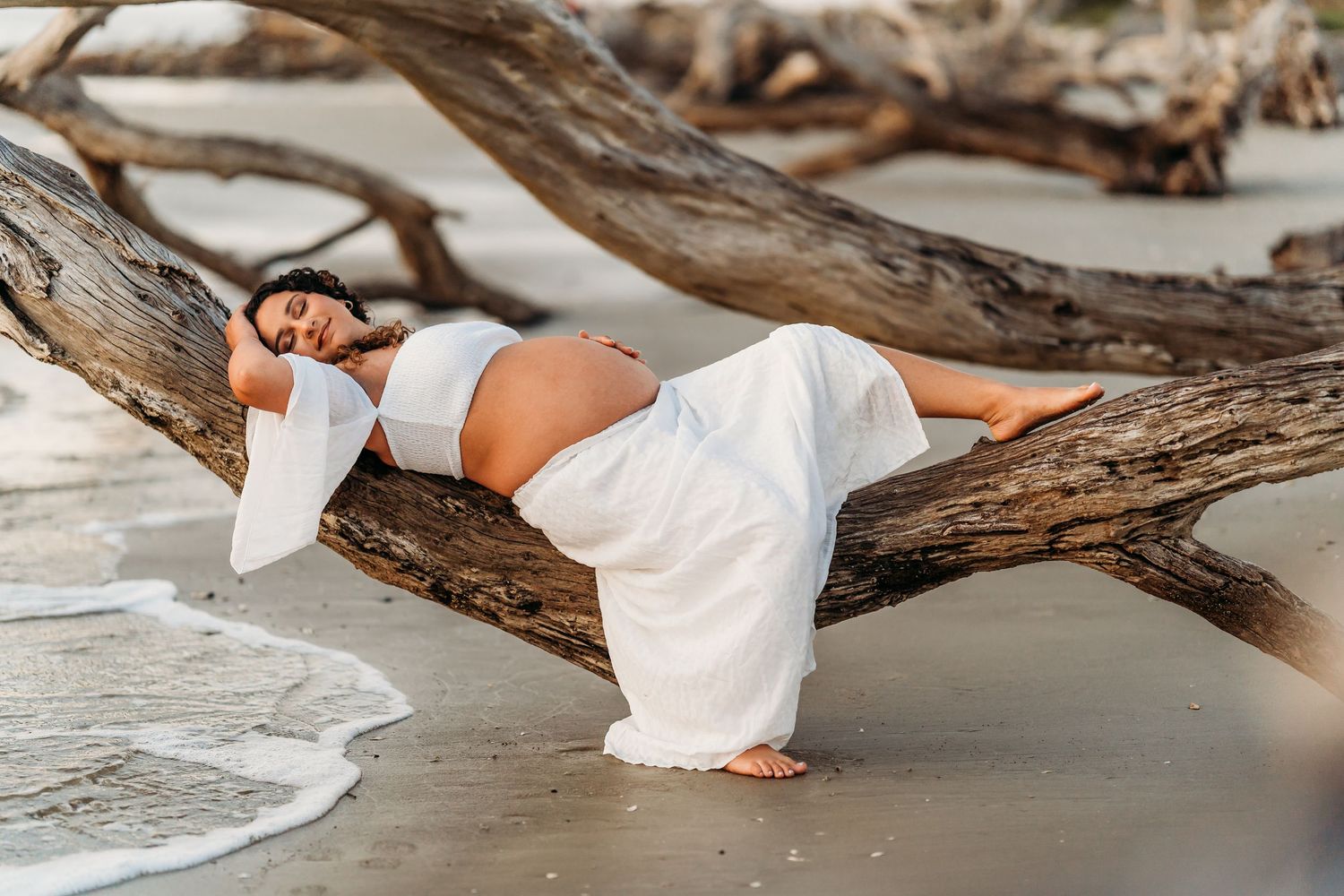Pregnant woman in white outfit poses gracefully on a driftwood log on a sandy beach.