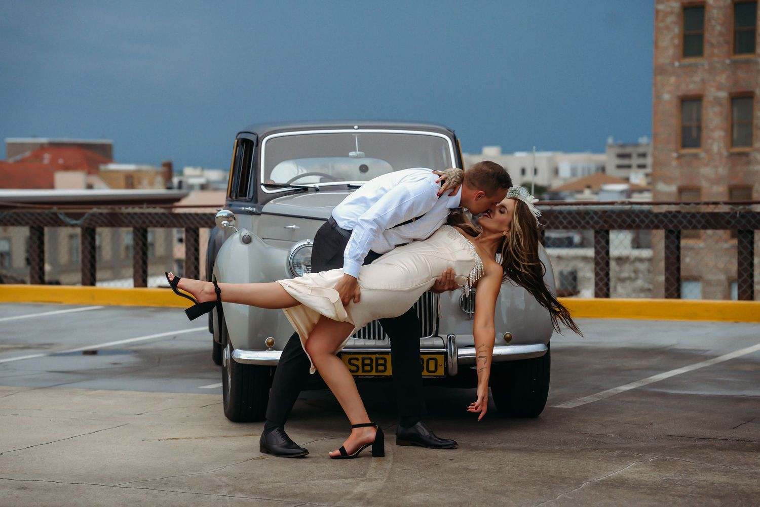 Couple shares a dramatic dip kiss in front of a vintage car on a rooftop.