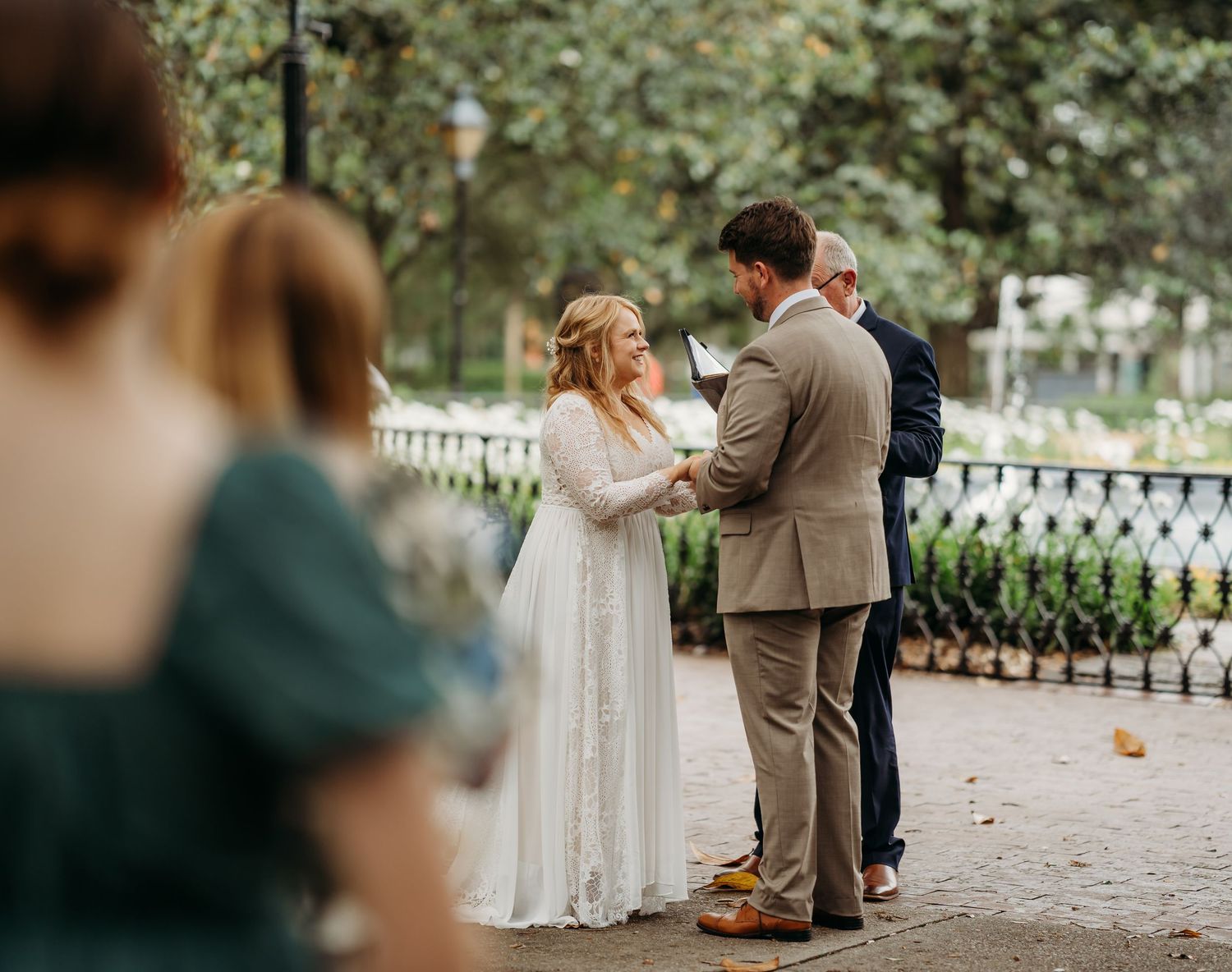 Bride in lace gown and groom in tan suit hold hands during outdoor wedding ceremony with guests watching.