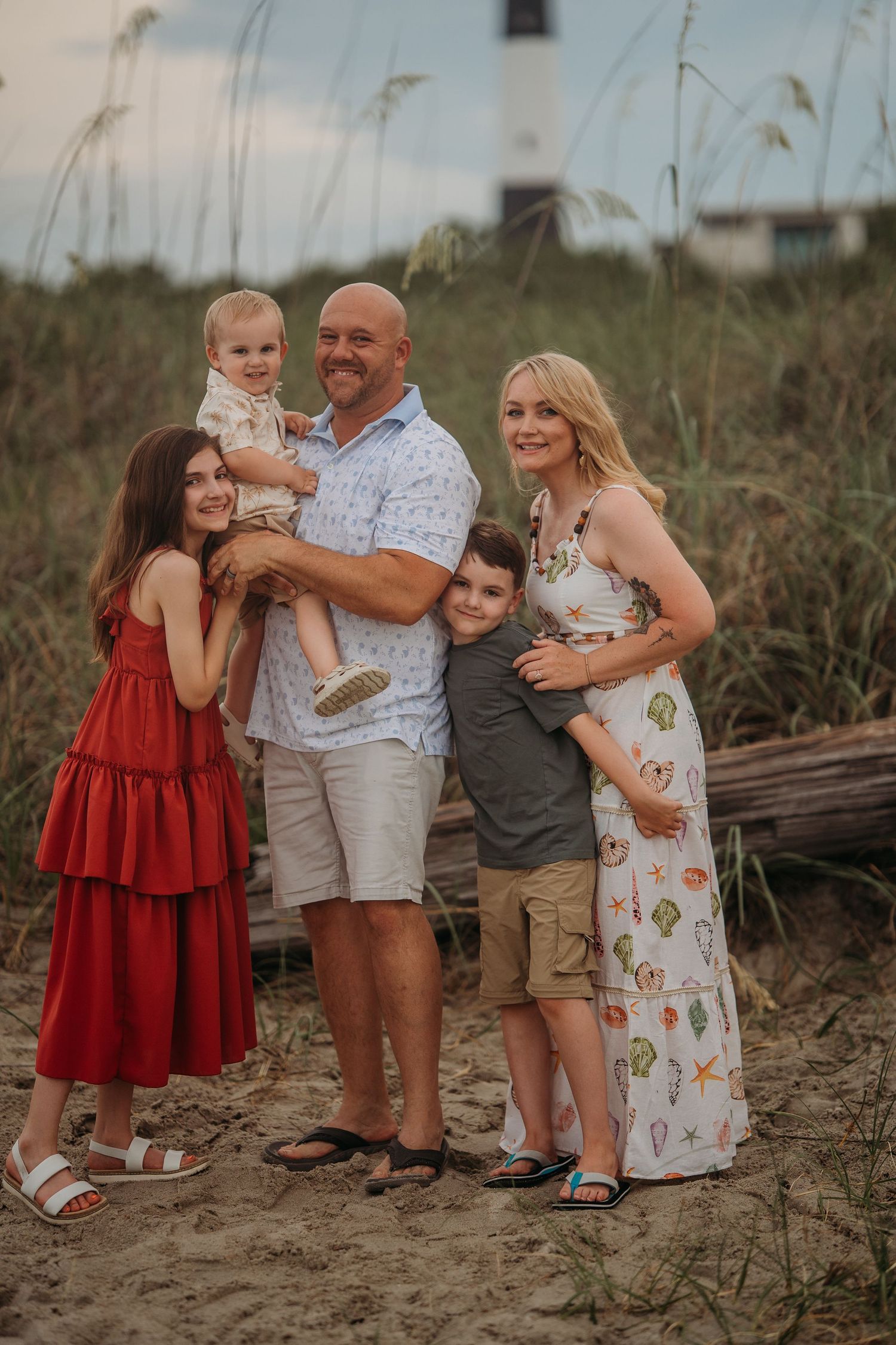 Family of five poses together near sand dunes and a lighthouse at the beach.