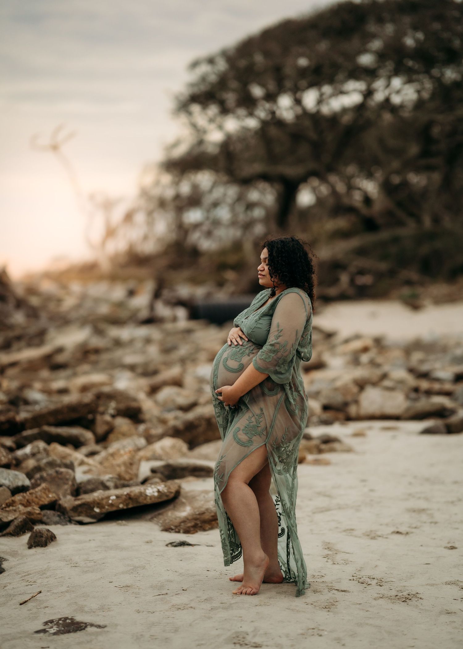 Pregnant woman in flowing green lace gown cradles her belly on a rocky beach at golden hour.