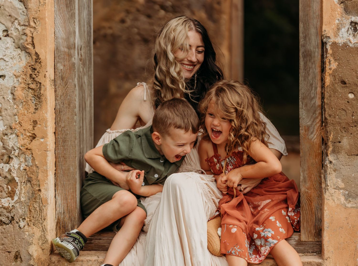 A joyful mother laughs with her two young children sitting in a rustic stone doorway.