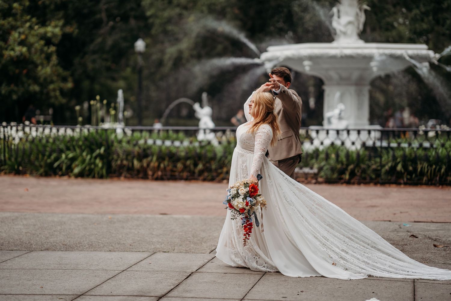 Groom embraces bride from behind near a grand white fountain, her long wedding dress train spread elegantly on the ground.