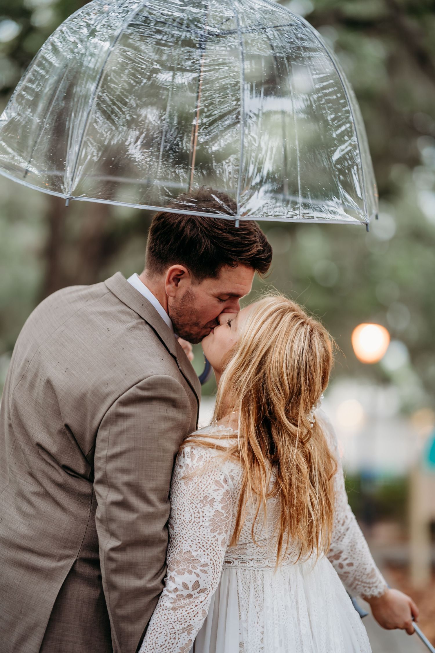 Bride and groom sharing a romantic kiss under a clear bubble umbrella on their rainy wedding day.