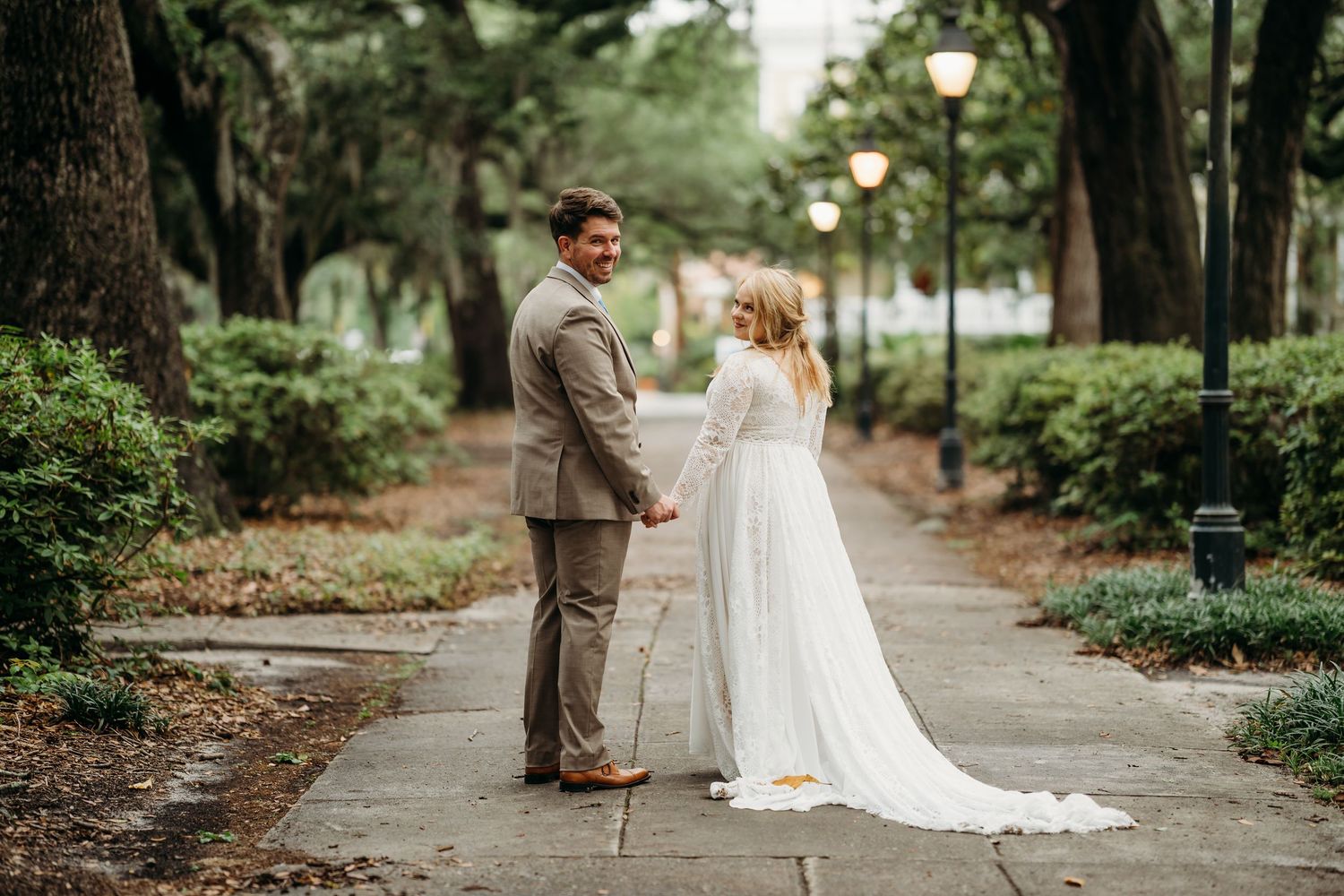Bride in long-sleeve white gown and groom in tan suit hold hands facing each other on a lush, lamp-lit garden pathway.