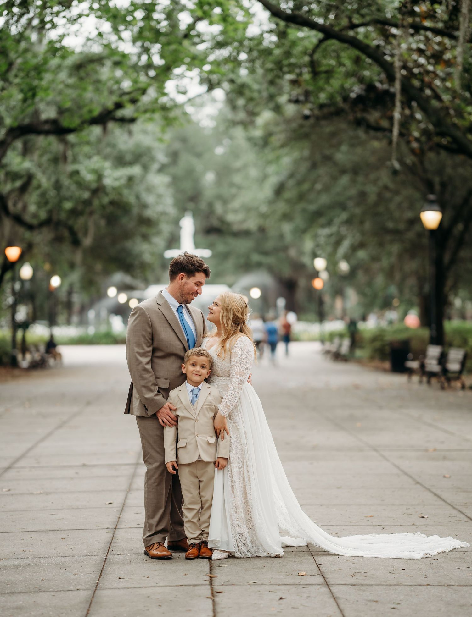 Wedding couple with young boy in suit share intimate moment on tree-lined park path with fountain.