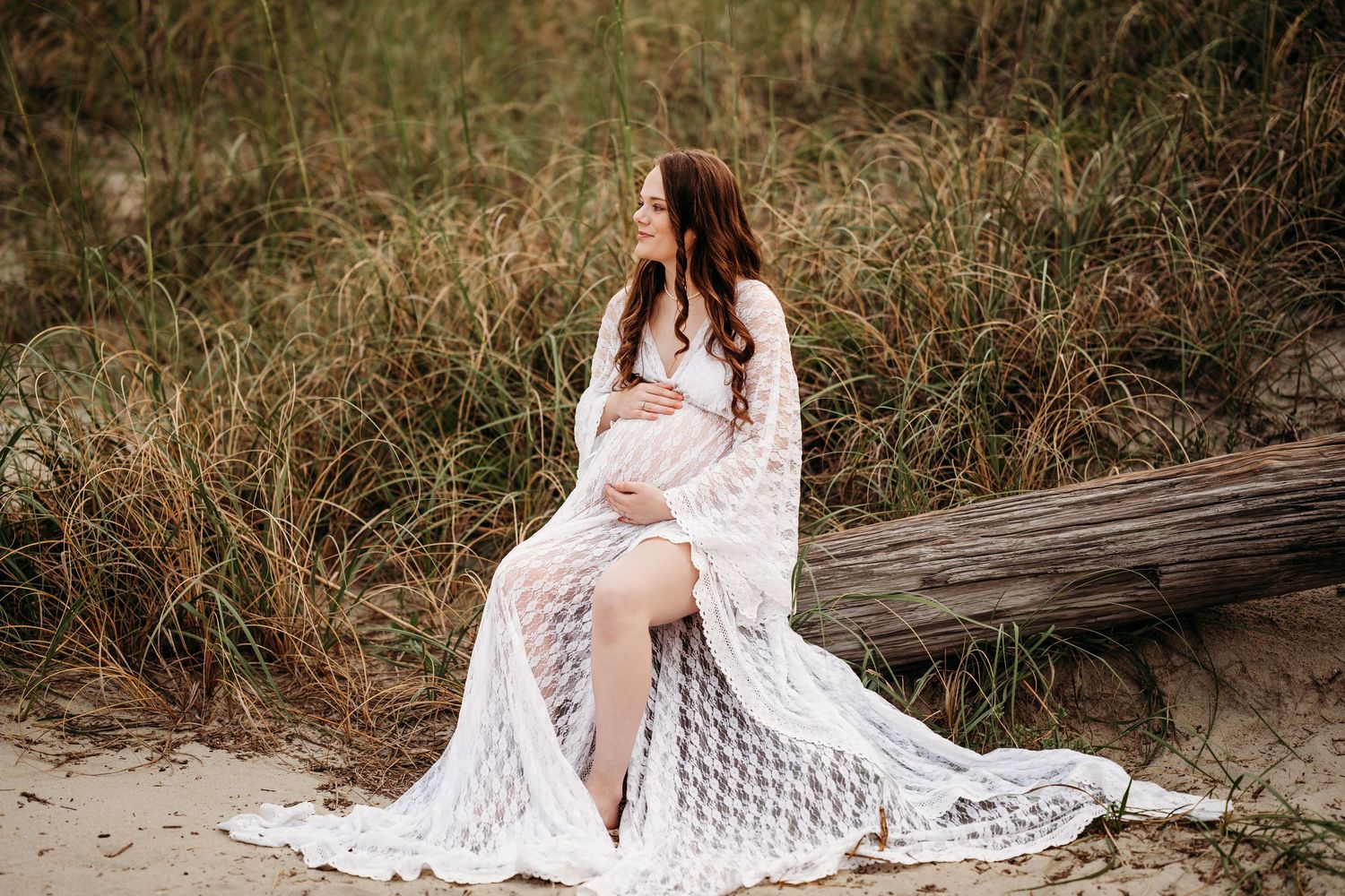 Expectant mother in white lace maternity gown sits on driftwood surrounded by beach dune grasses.