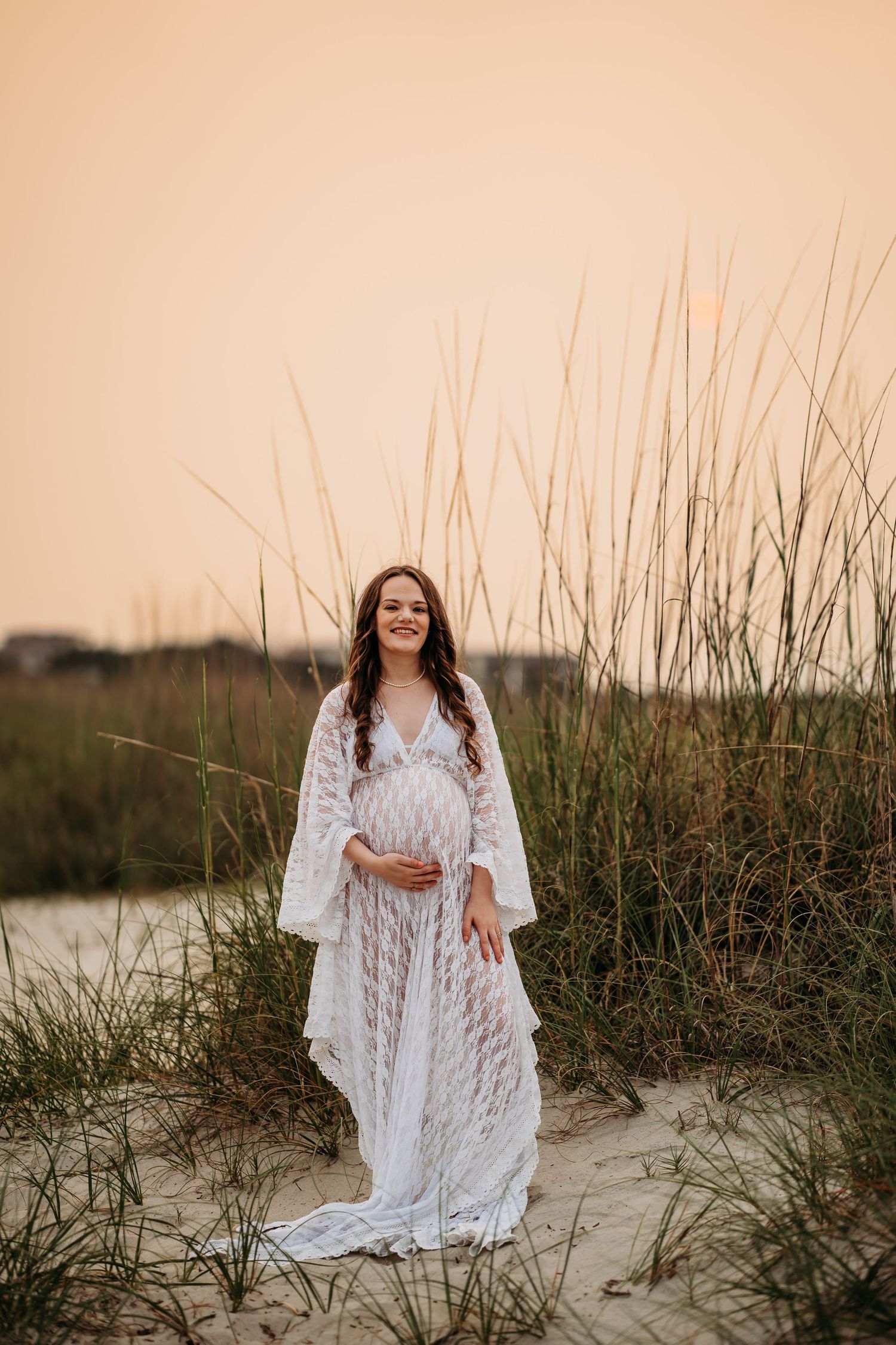 Pregnant woman in flowing white lace dress standing among beach dunes at warm sunset.