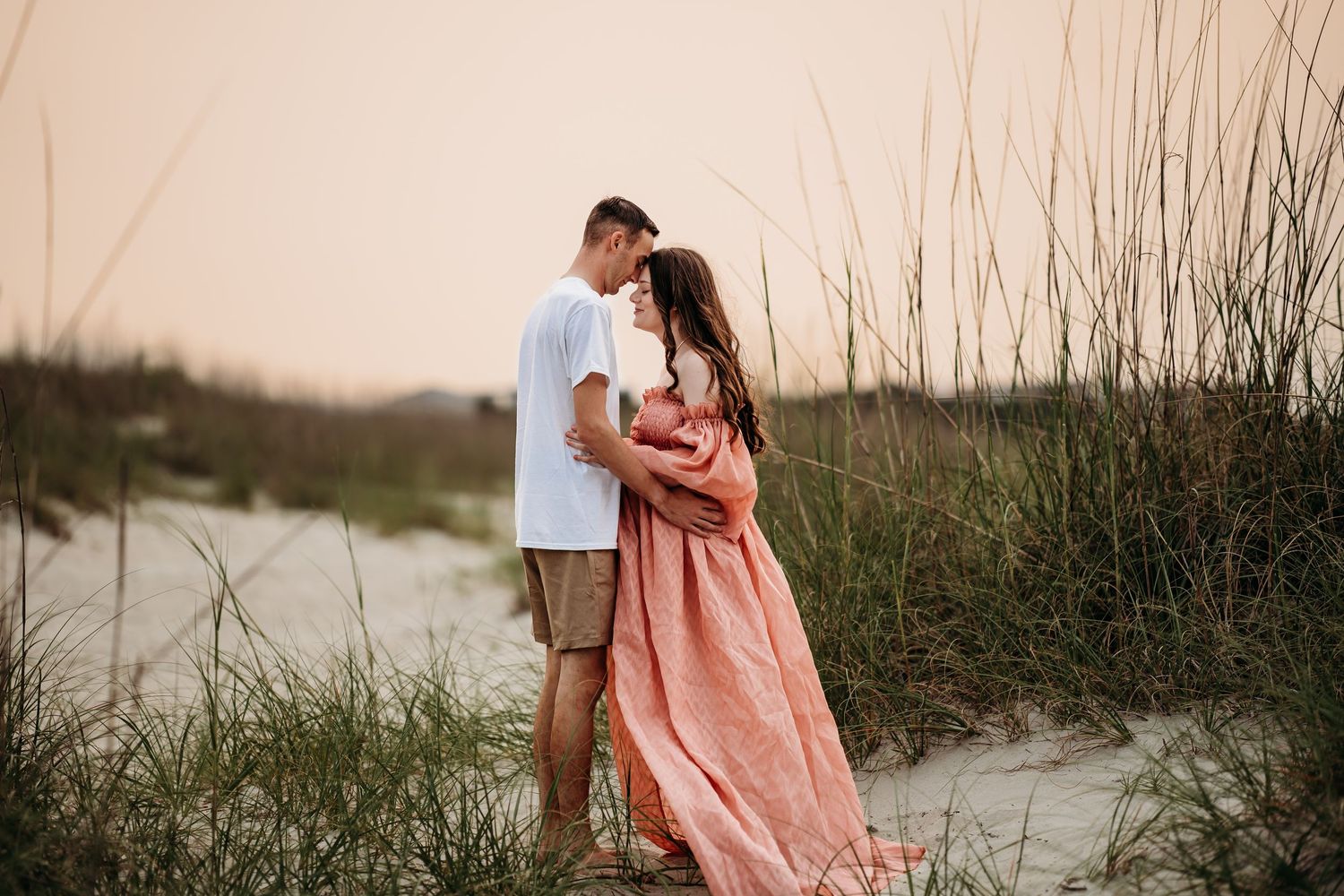 Expecting couple sharing tender moment on sandy beach dunes at sunset, woman in coral maternity dress.