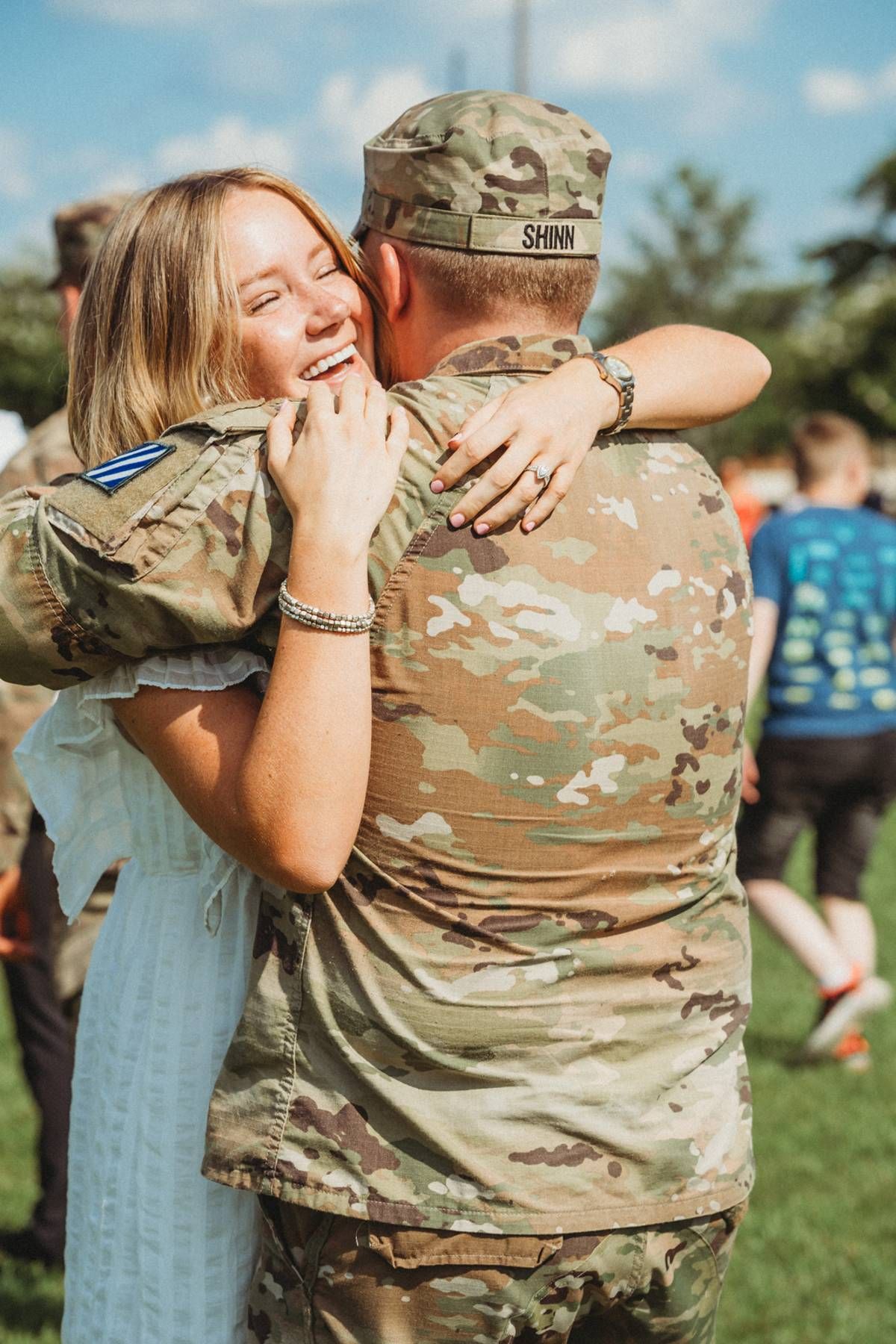 Smiling woman joyfully embraces a returning soldier in camouflage uniform outdoors.