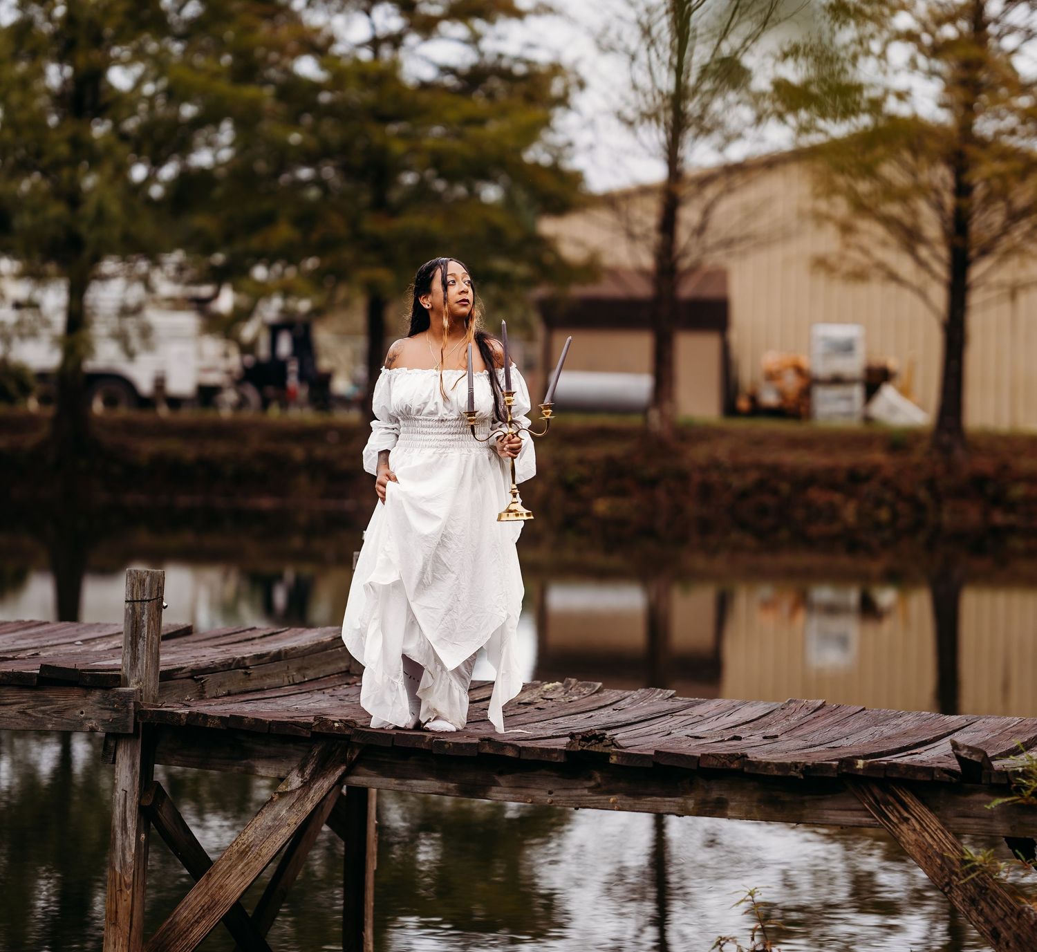 Woman in white off-shoulder dress walks along wooden bridge over reflective pond holding scales.