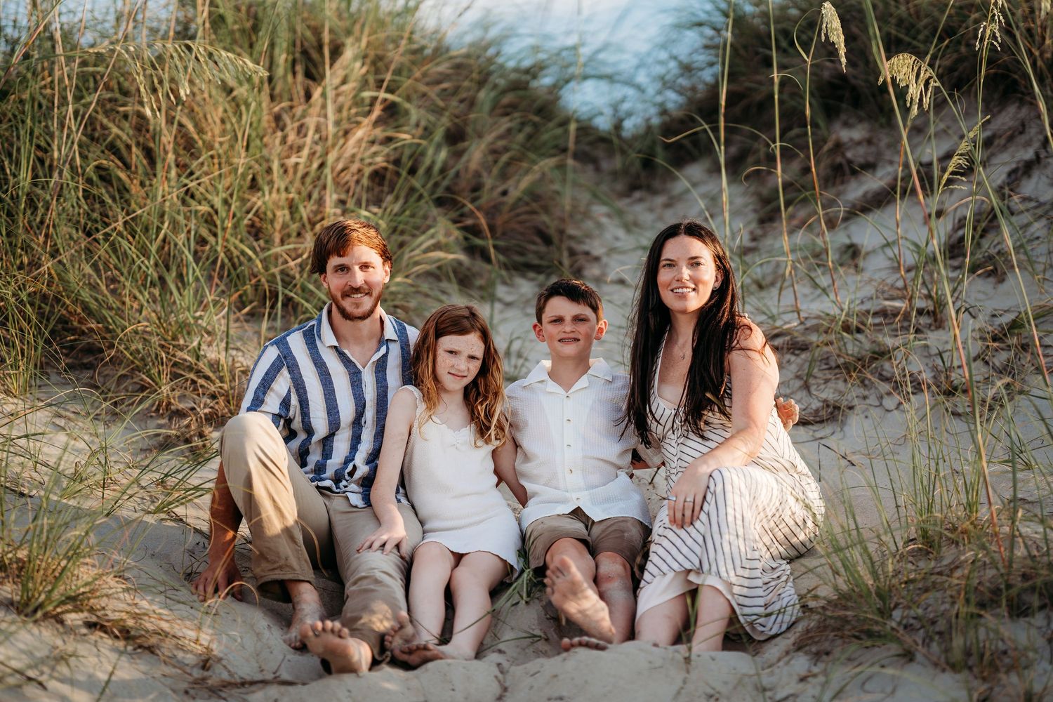 Family of four smiling together on sandy beach dunes with sea grass, casual coastal photo session.