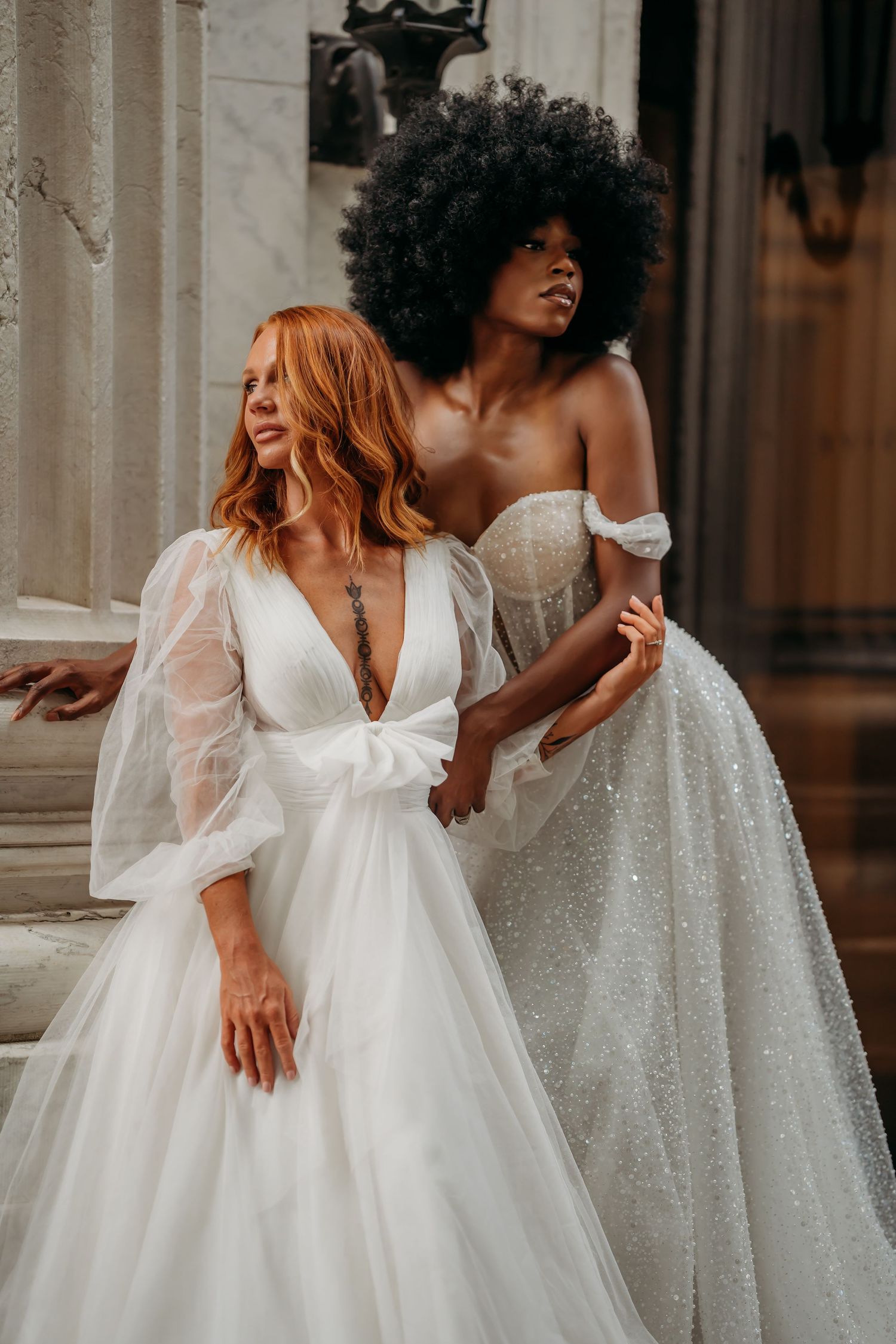Two brides in elegant white wedding gowns, one in sheer sleeves and one in off-shoulder sparkle, pose on stone steps.