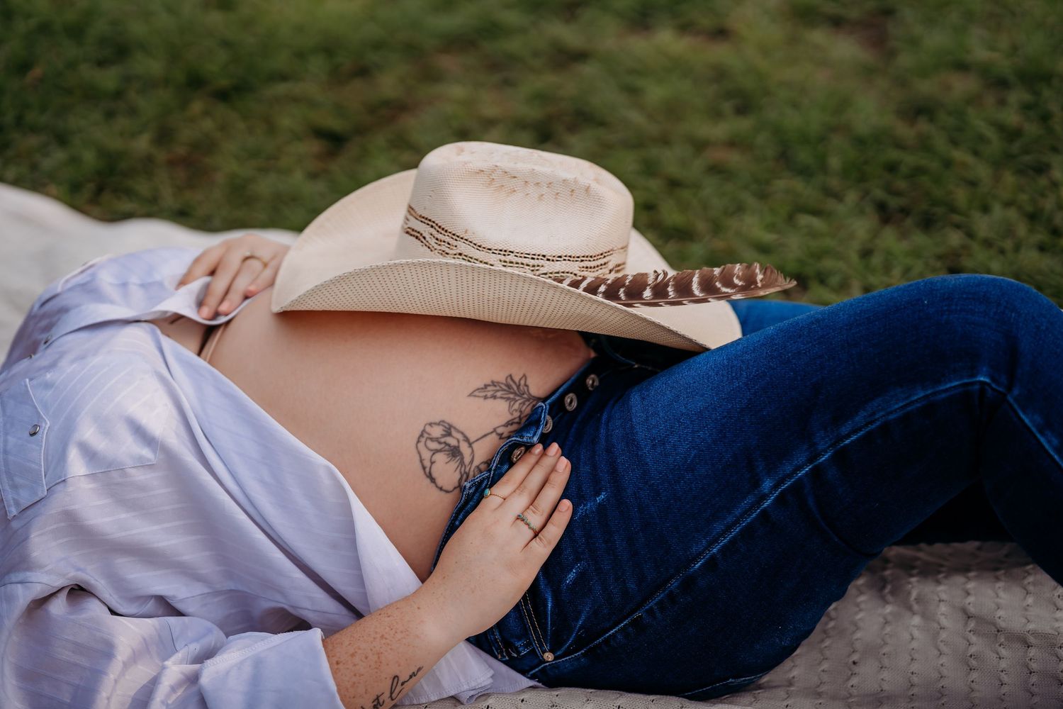 Pregnant woman in cowboy hat and jeans lying down, showing tattooed baby bump outdoors.